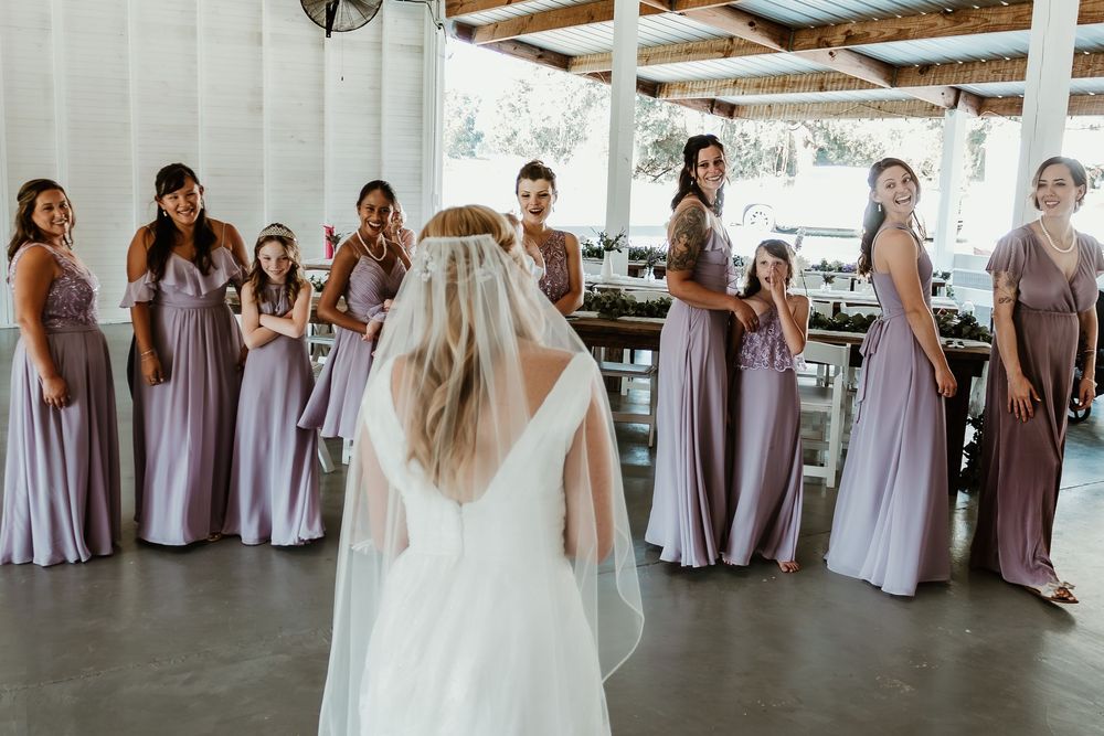 A bride in a backless white gown faces her bridesmaids wearing coordinated purple dresses.