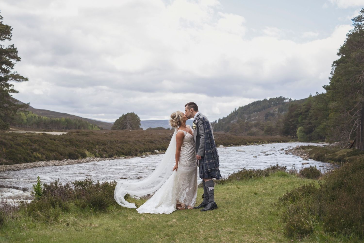 A bride and groom kissing in a Scottish Highland landscape beside a river wearing a kilt and wedding dress.