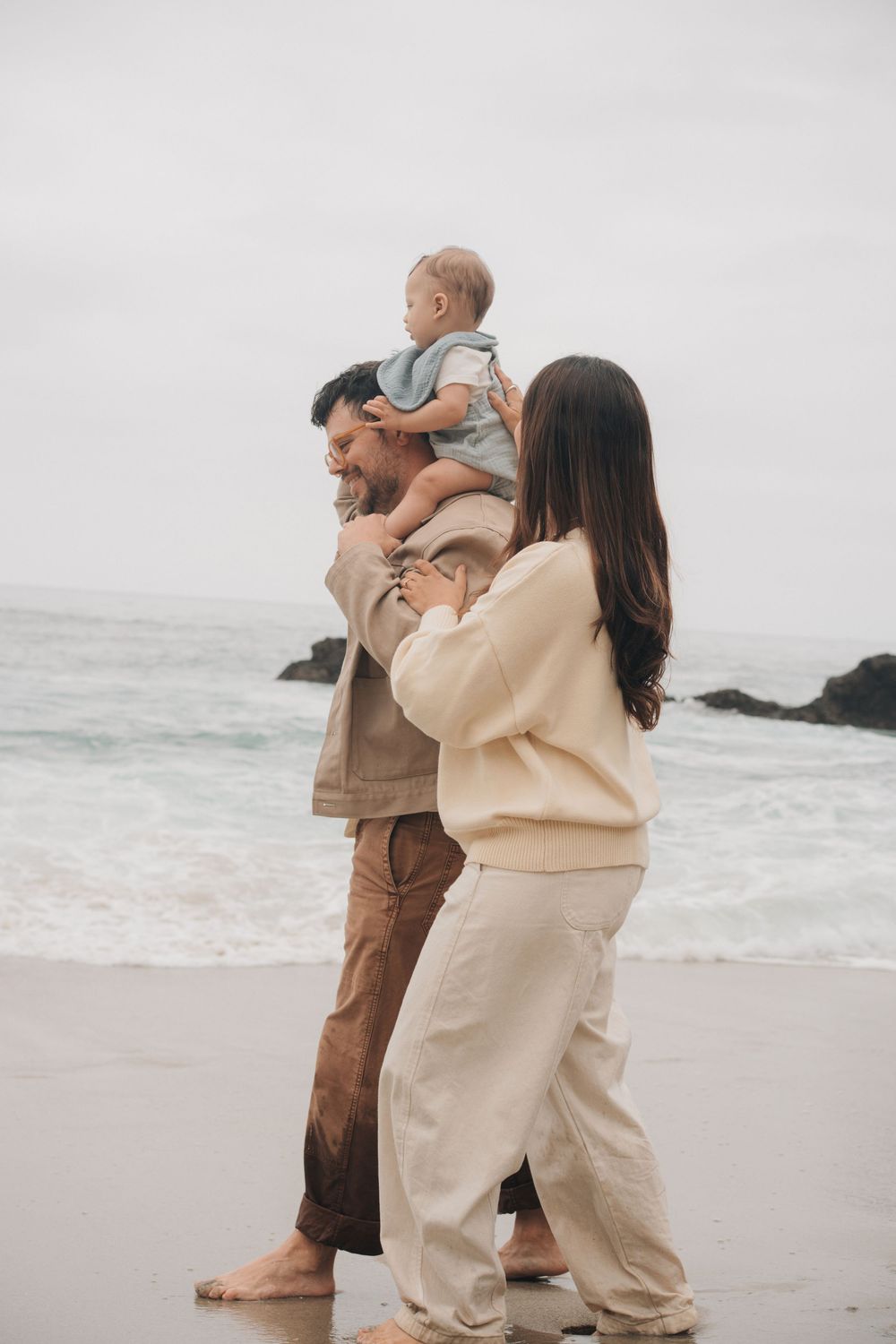 A couple in casual beige clothing walk together along a misty beach shoreline on an overcast day.