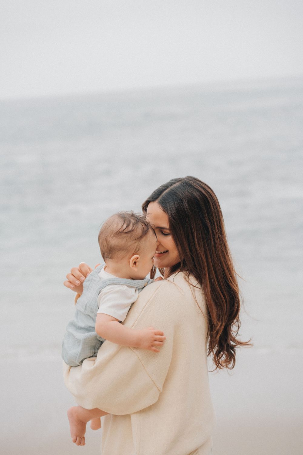 A tender moment captured between mother and baby in soft beige tones against a misty beach backdrop.