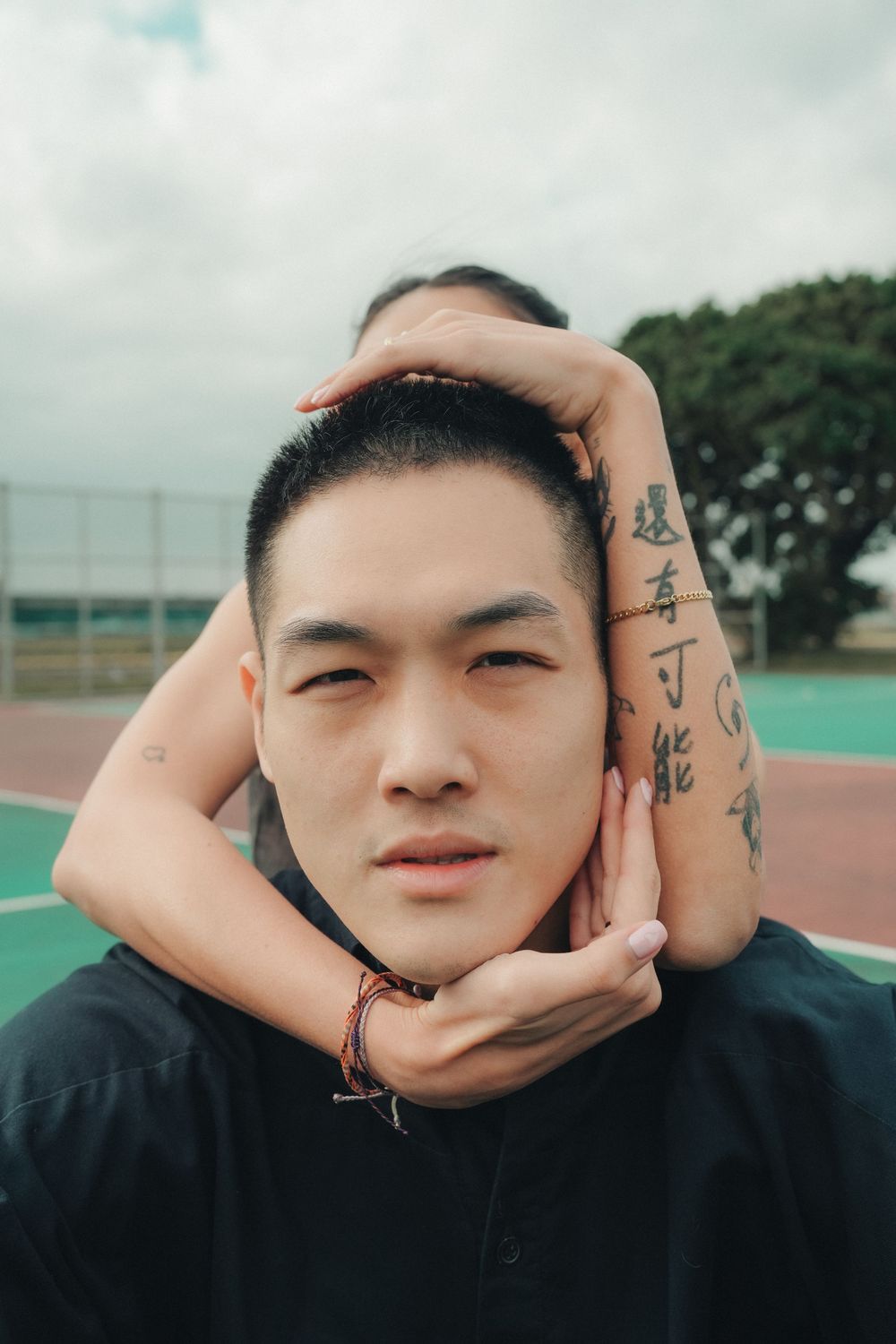Person with tattoos on their hands frames their face while posing outdoors on a sports field.
