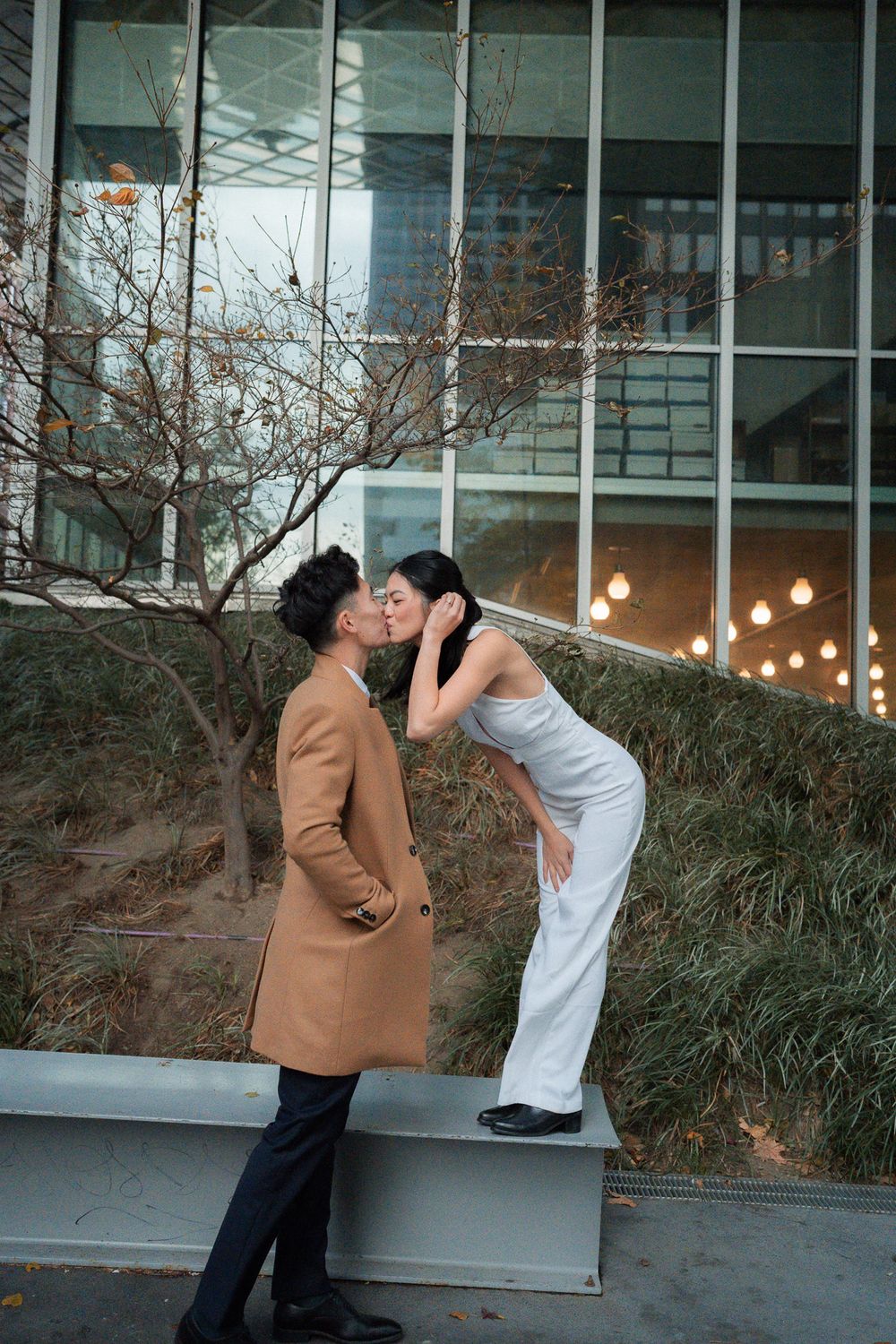 A couple shares a romantic moment outdoors near a modern building with glass windows and bare winter trees.
