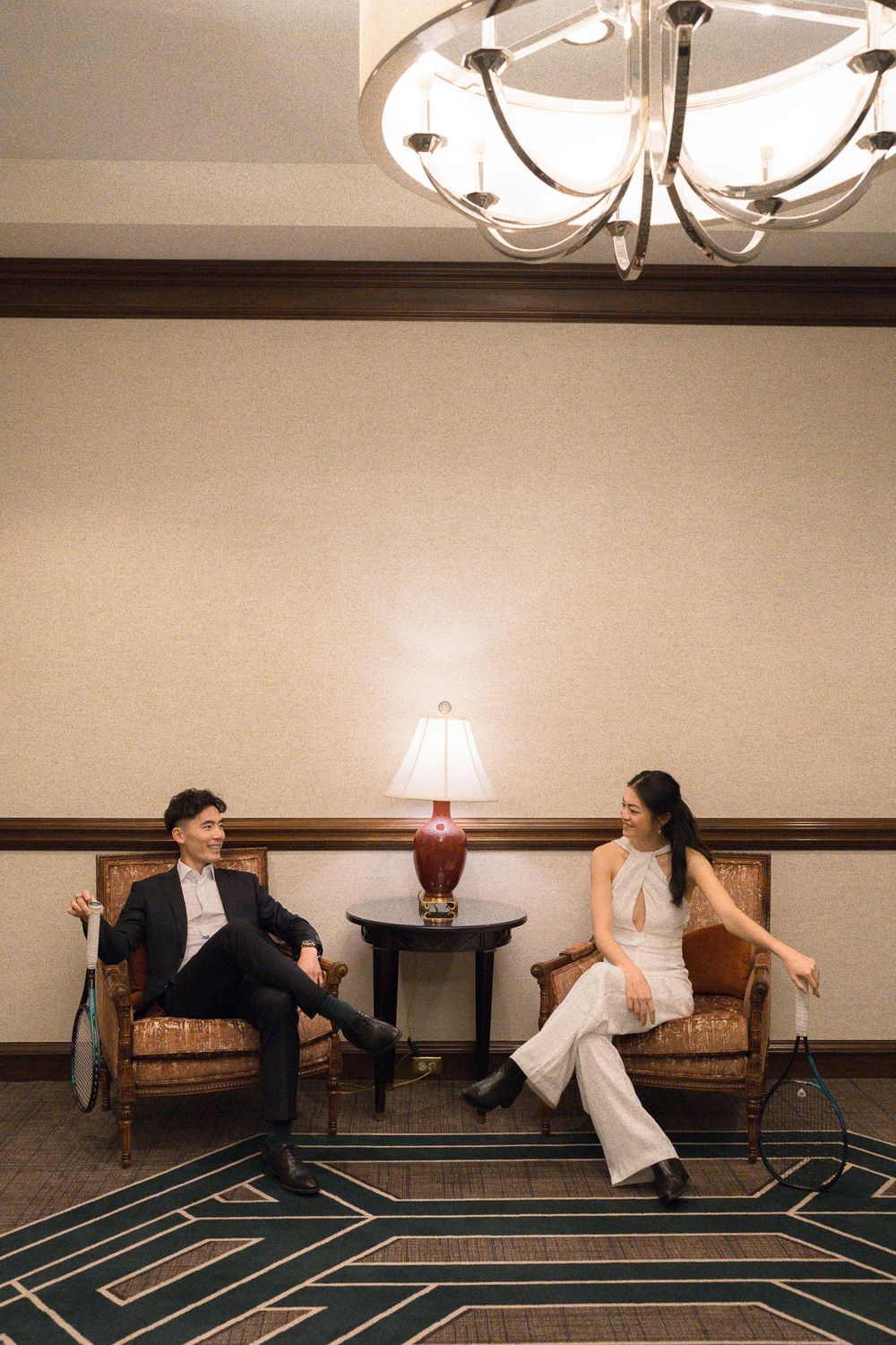 Two people sit in elegant armchairs having a conversation beneath a modern chandelier in an upscale hotel setting.