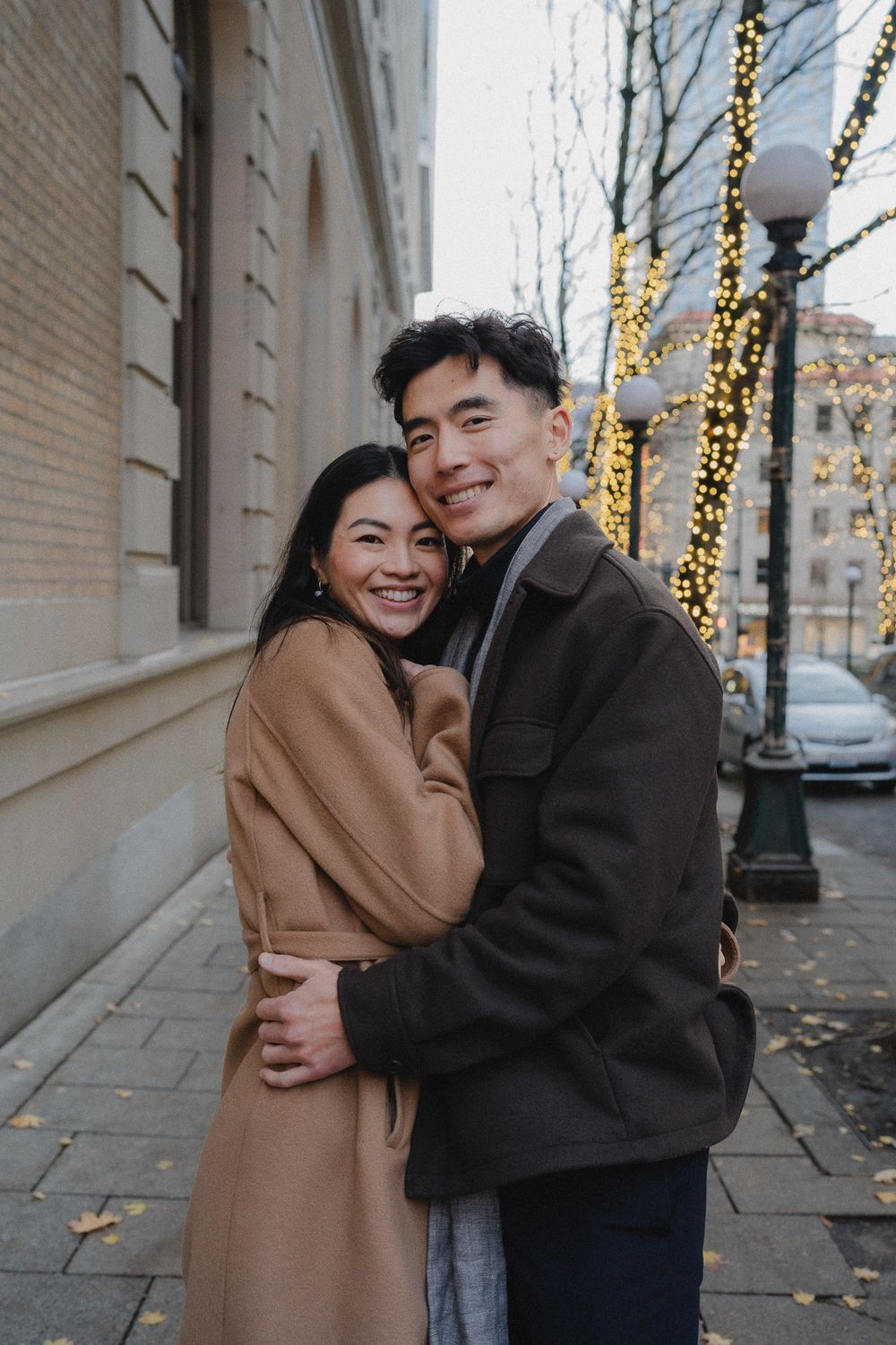 A couple shares joyful embraces along a brick building wall decorated with twinkling string lights.