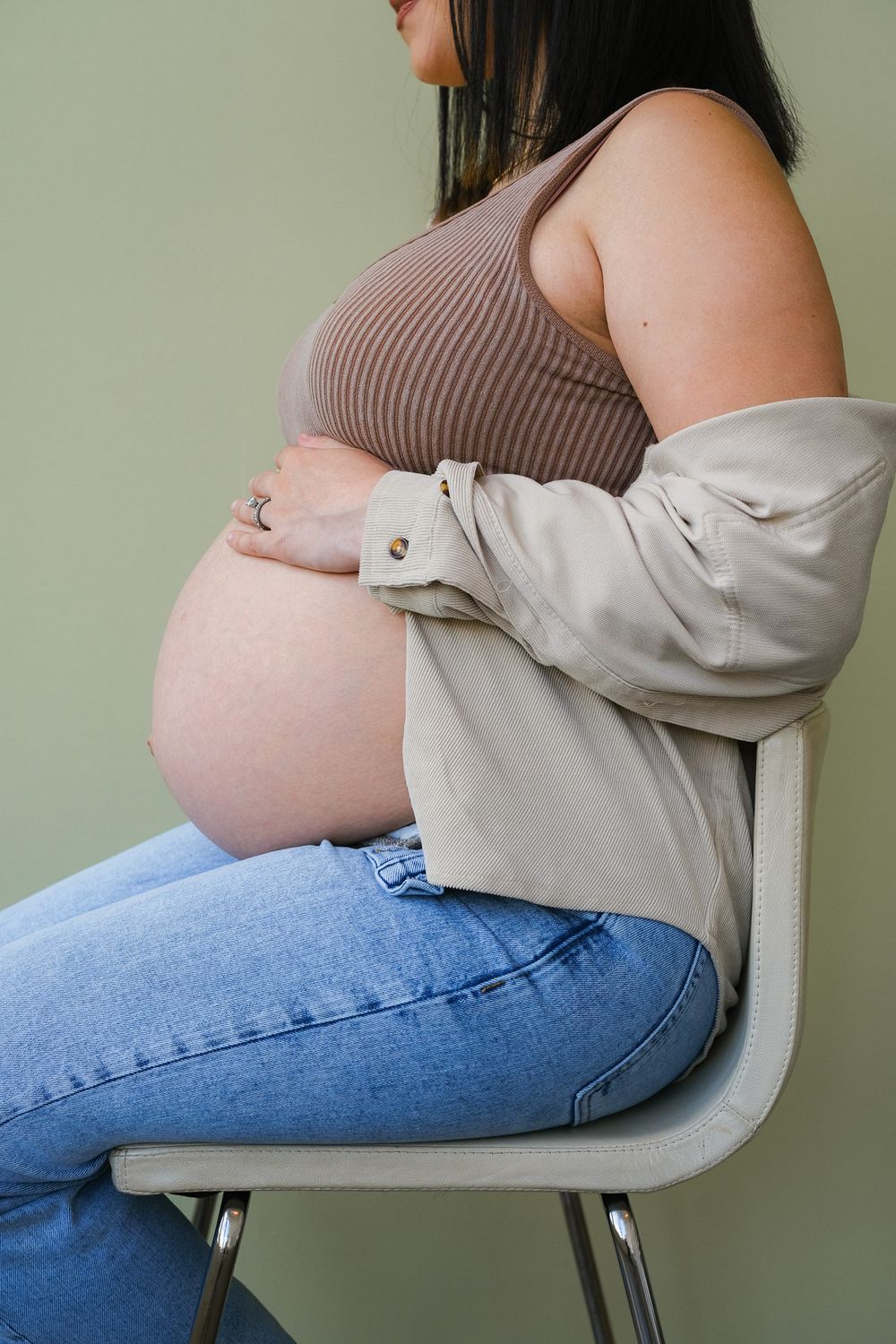 Side view of an expectant mother wearing a ribbed tank top, beige cardigan and jeans while sitting on a chair.
