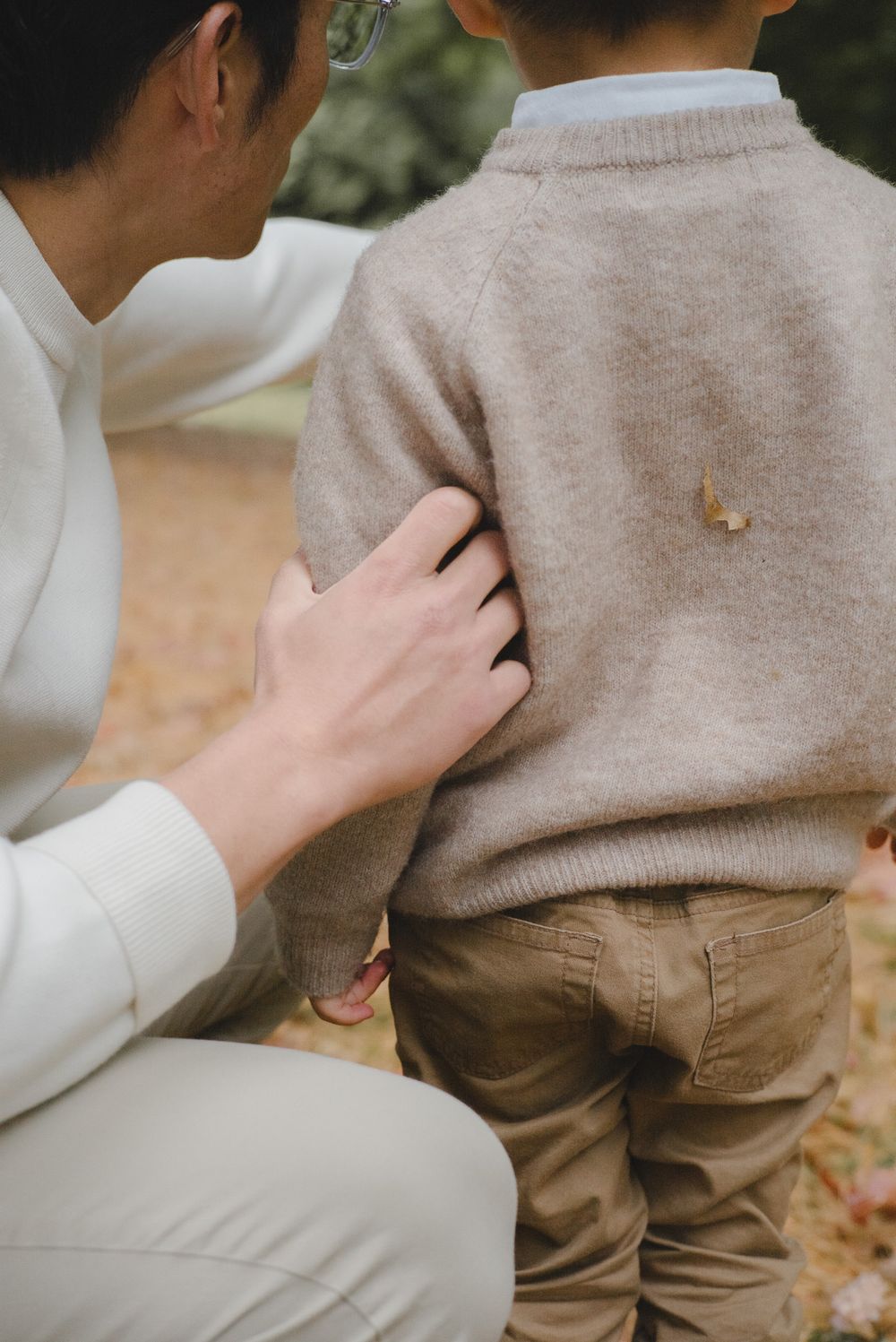 A person in a beige sweater and brown pants sits while another person's hand rests gently on their shoulder.