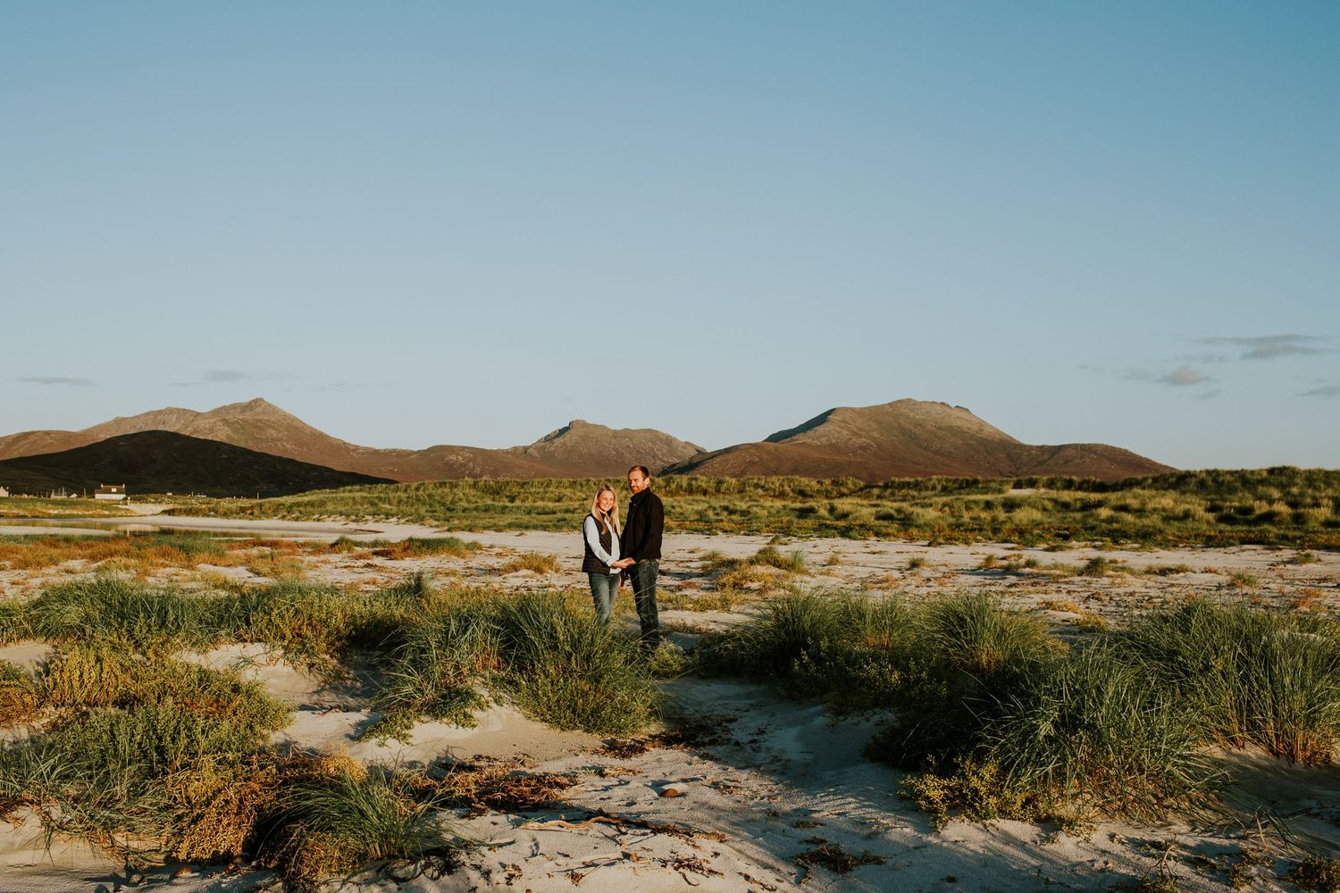A couple stands on a beach with South Uist hills silhouetted against a clear blue sky.