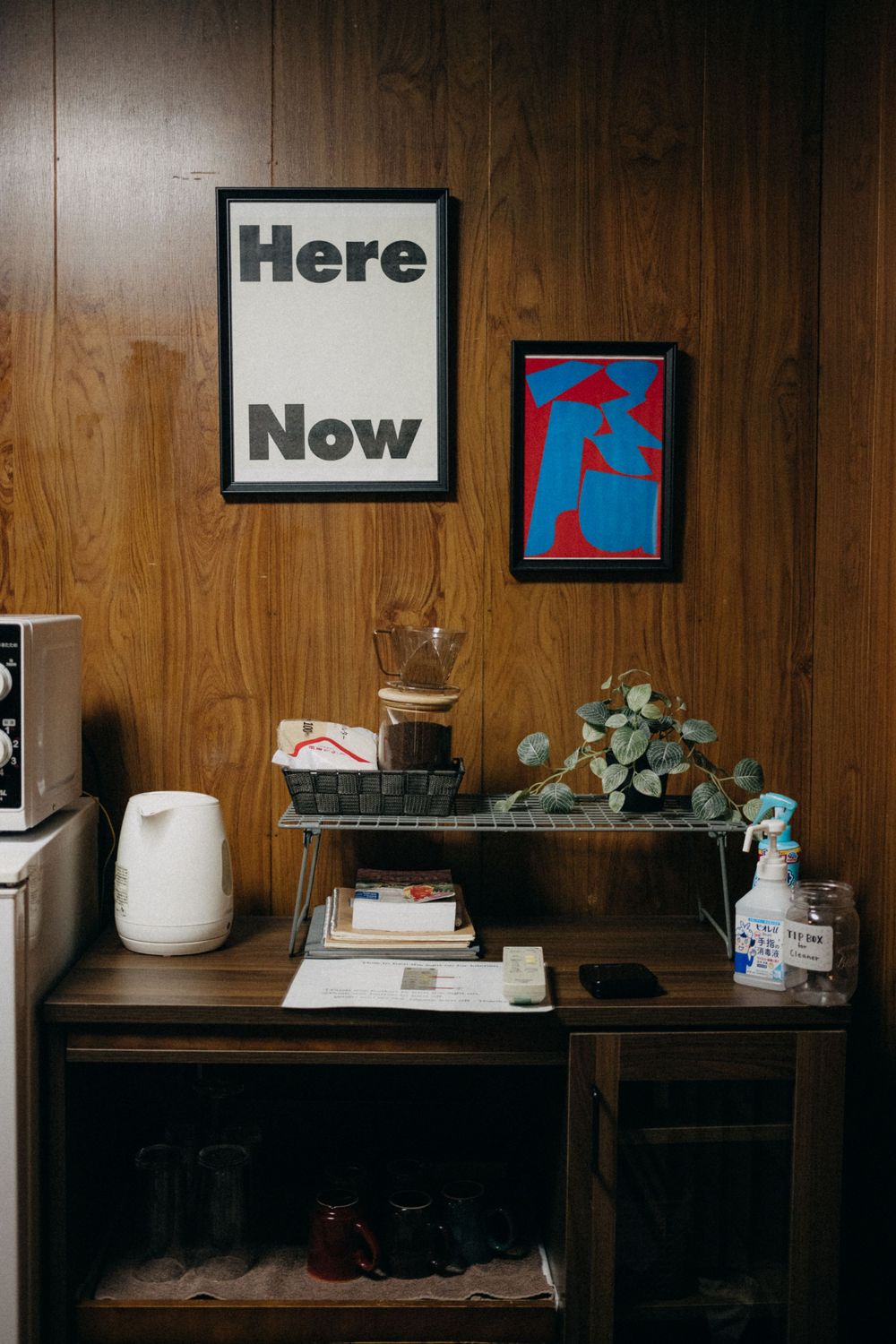 Mid-century modern shelf styling with 'Here Now' artwork against wood-paneled wall.