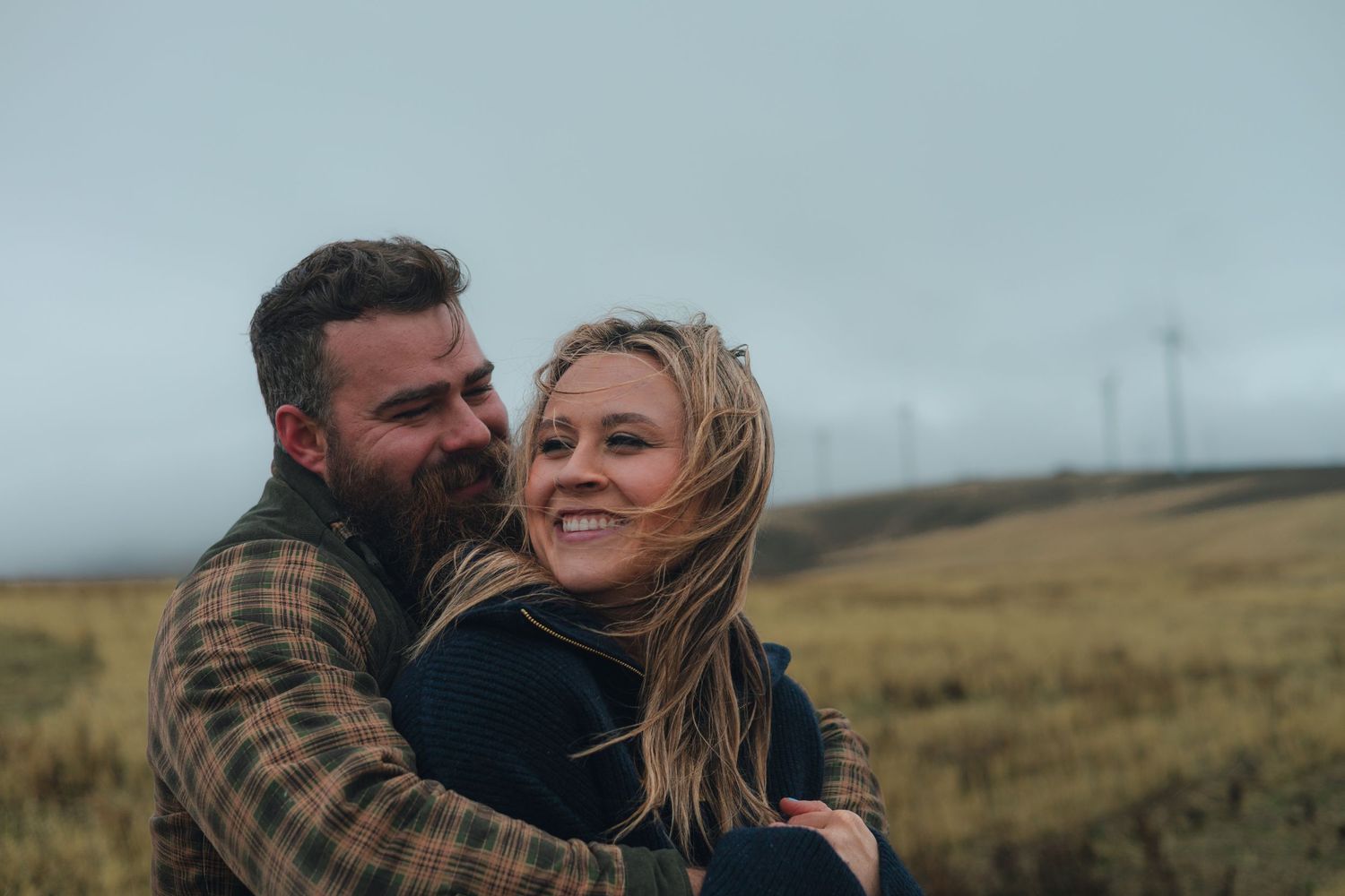 A couple embraces on a windswept hill with wind turbines visible in the misty background.