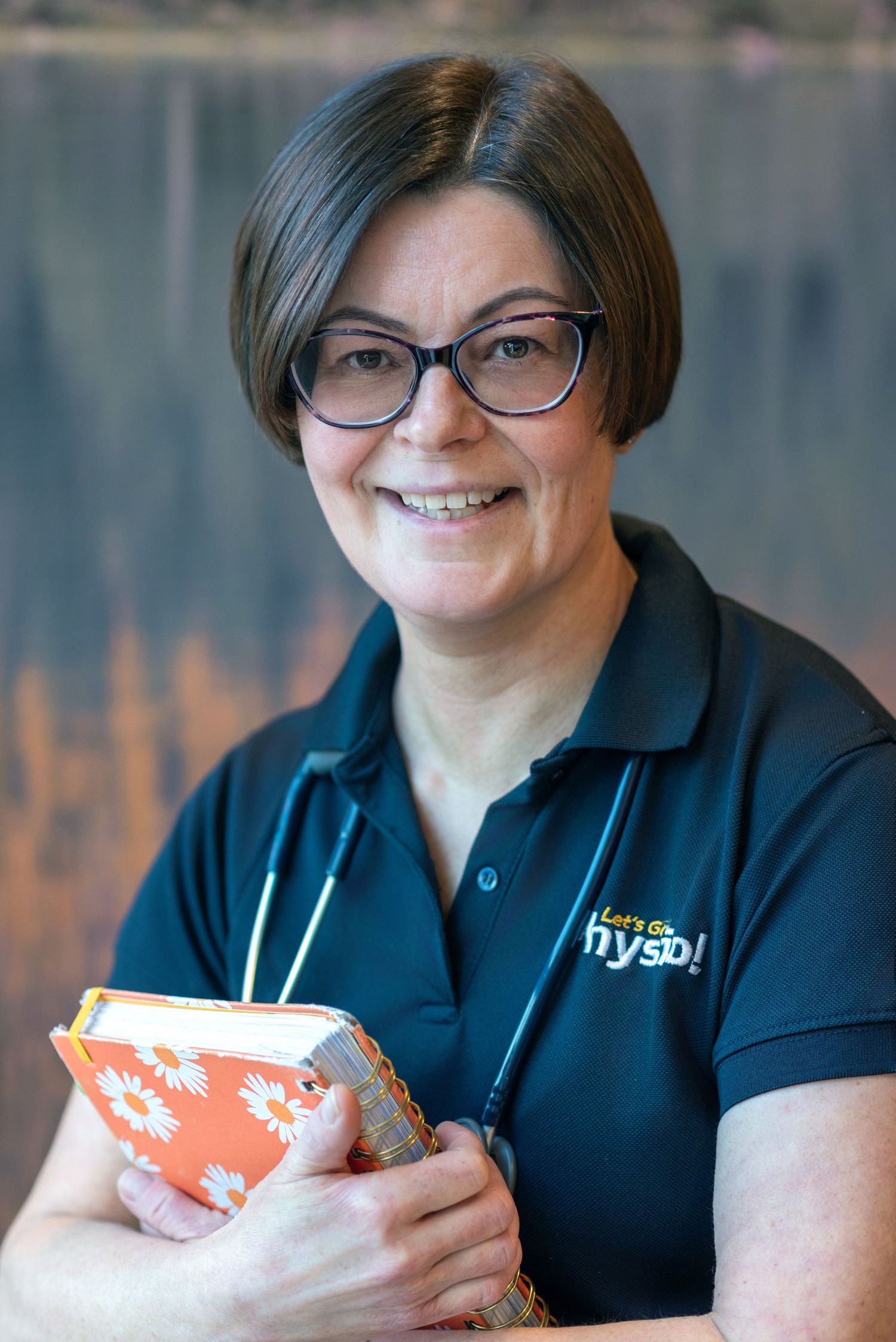 Healthcare worker in navy blue polo shirt with stethoscope smiling while holding an orange and white box.