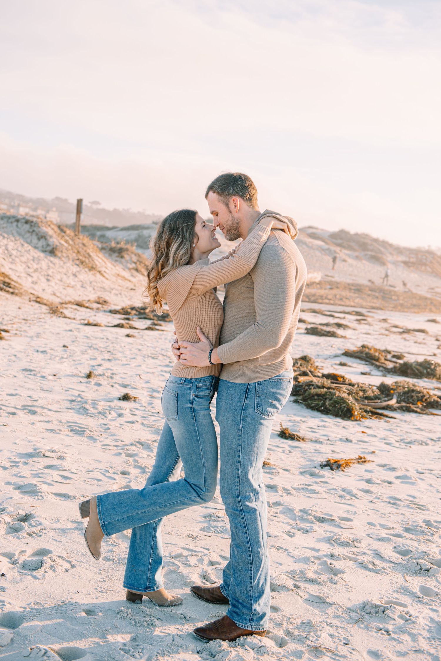 A couple embraces romantically on a sandy beach during winter with snow-covered hills in the background.