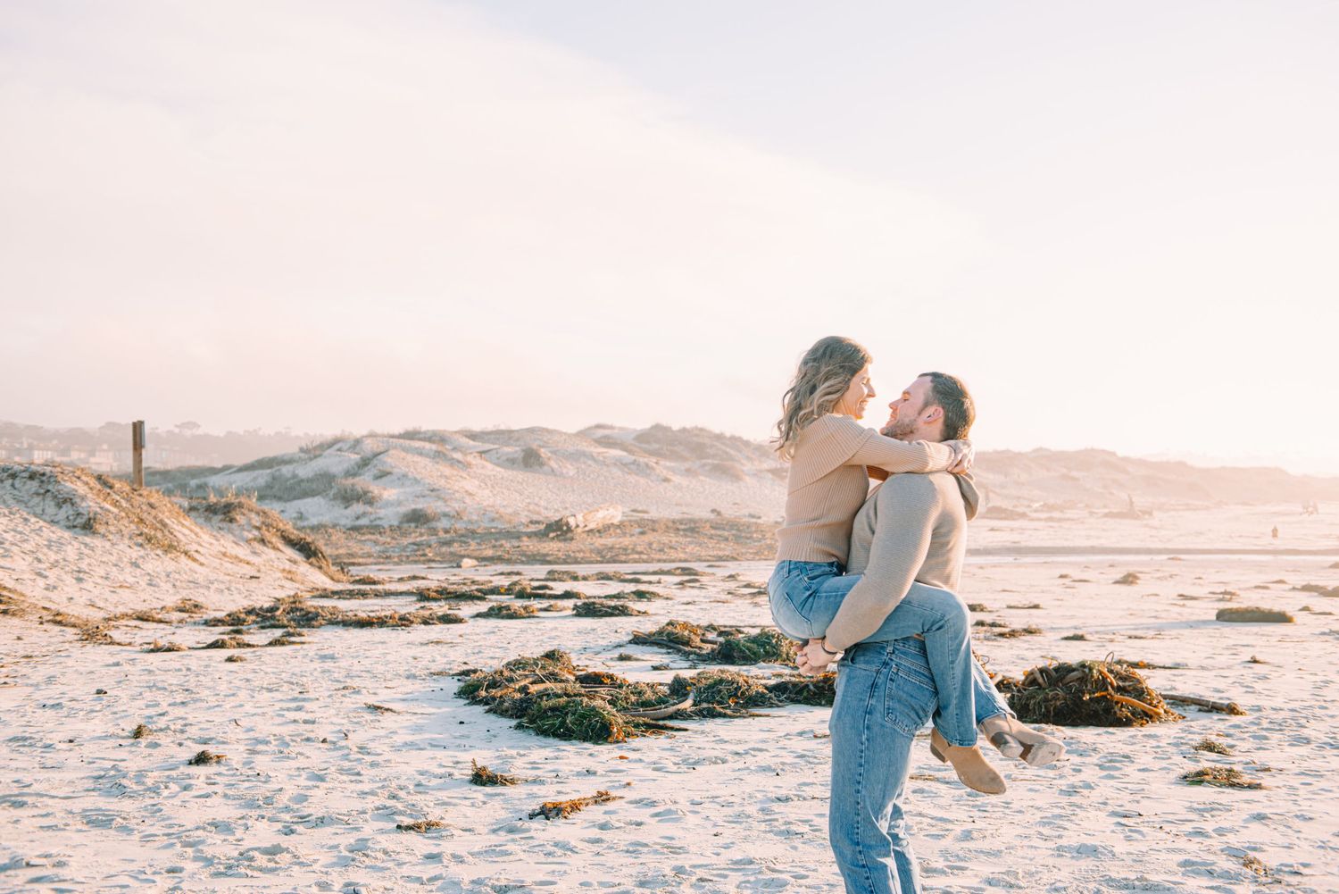 Two people share a tender moment on a sandy beach with hills in the background during golden afternoon light.