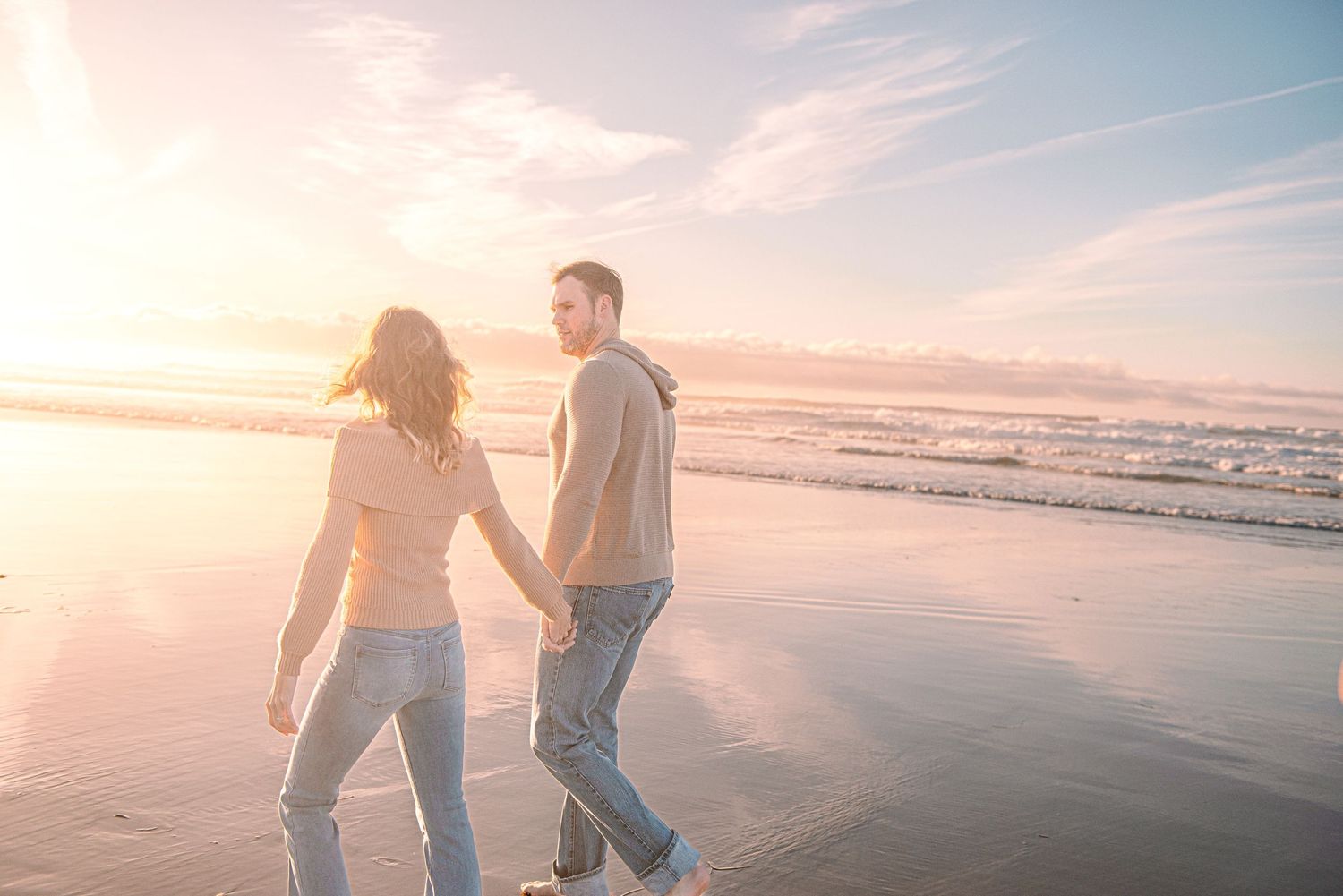 A couple walks hand in hand along a misty beach shore during golden sunset.