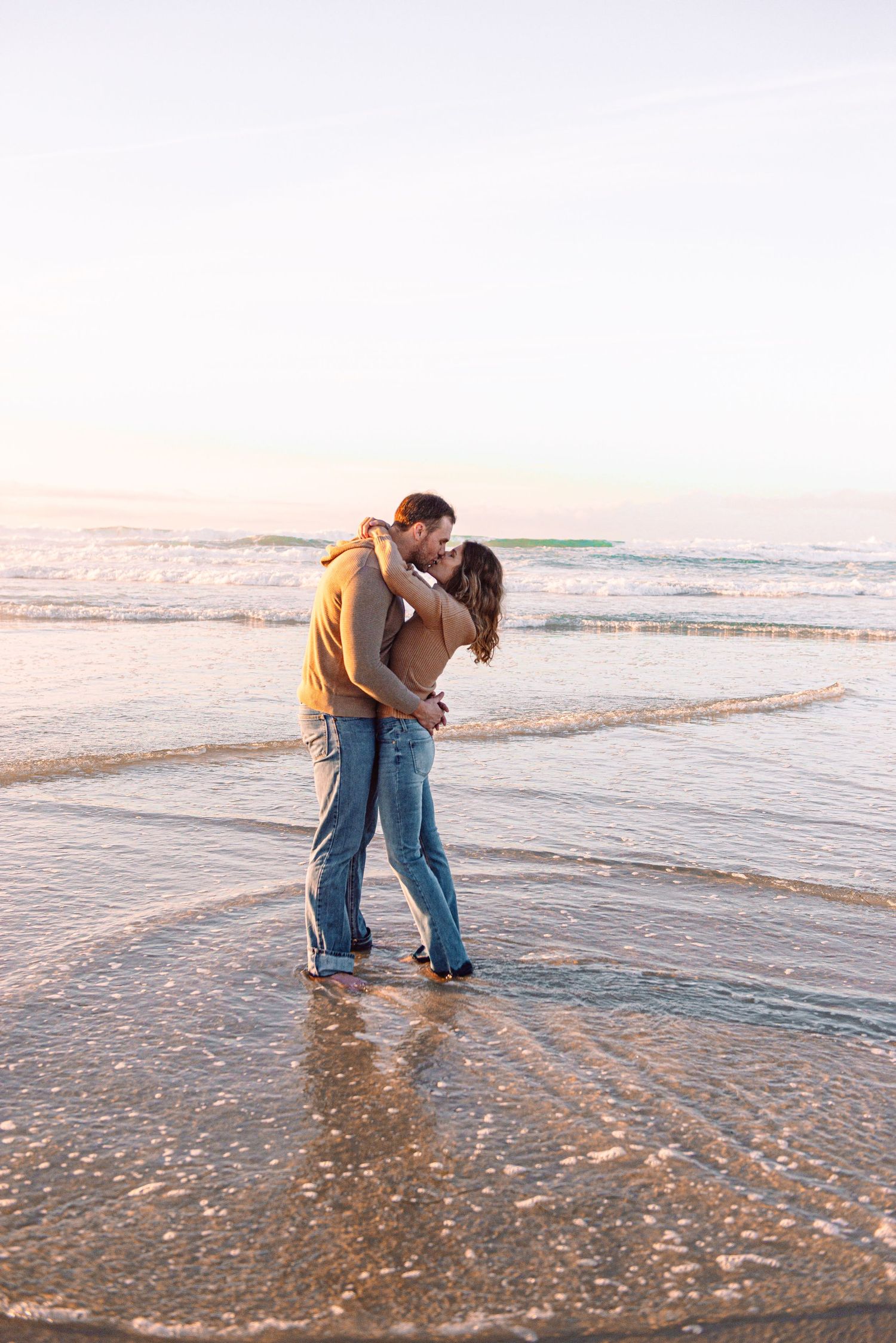 A couple shares a romantic kiss while standing in shallow ocean waves at sunset on a beach.