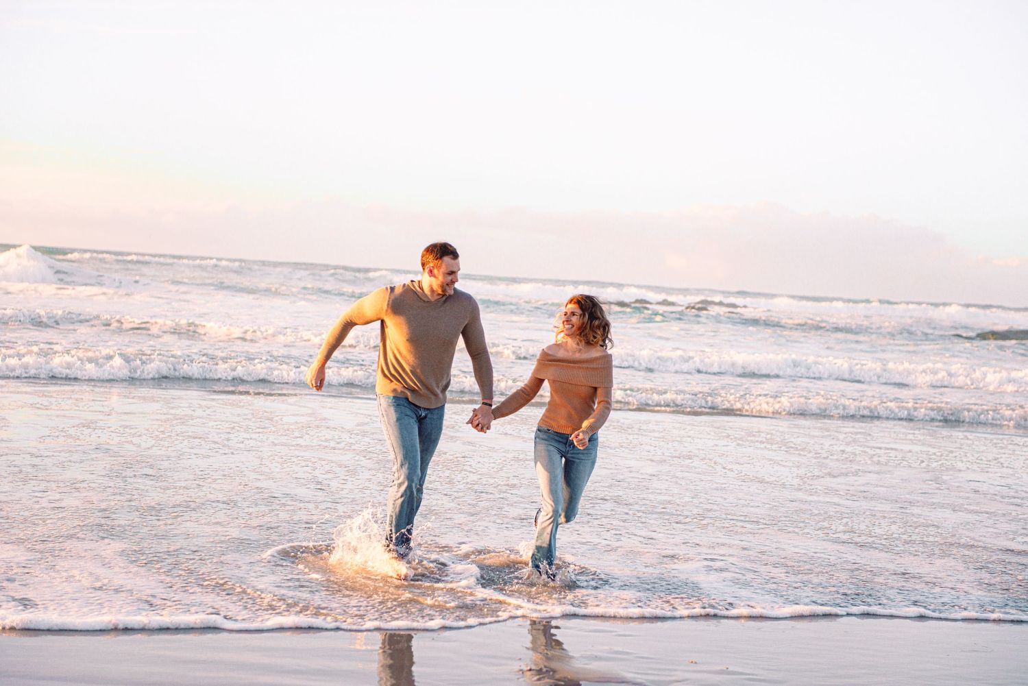 Two people holding hands and running along the beach at sunset as waves splash around their feet.