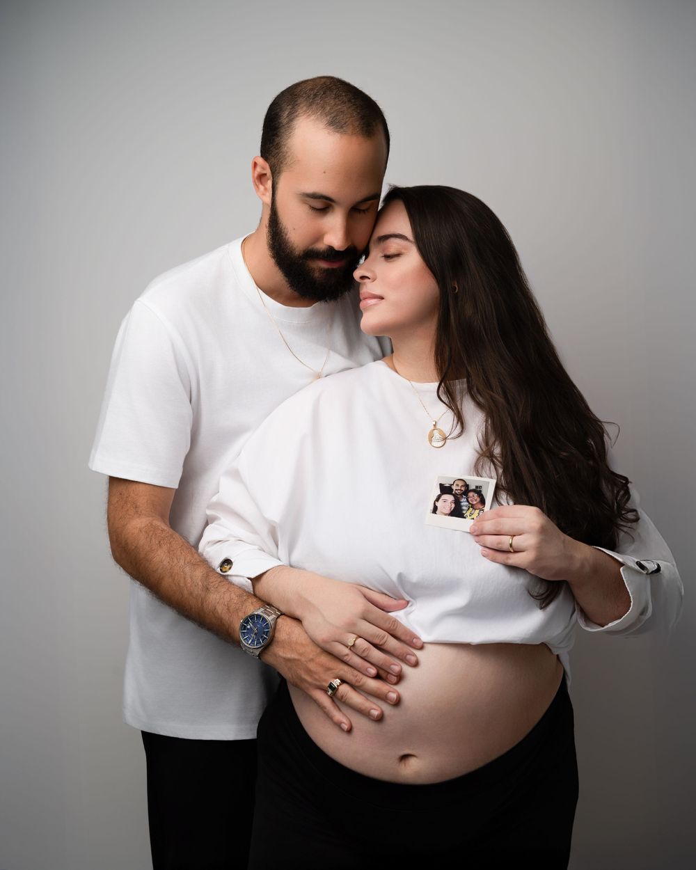 Expecting couple in white outfits holding ultrasound photo over baby bump in maternity shoot.