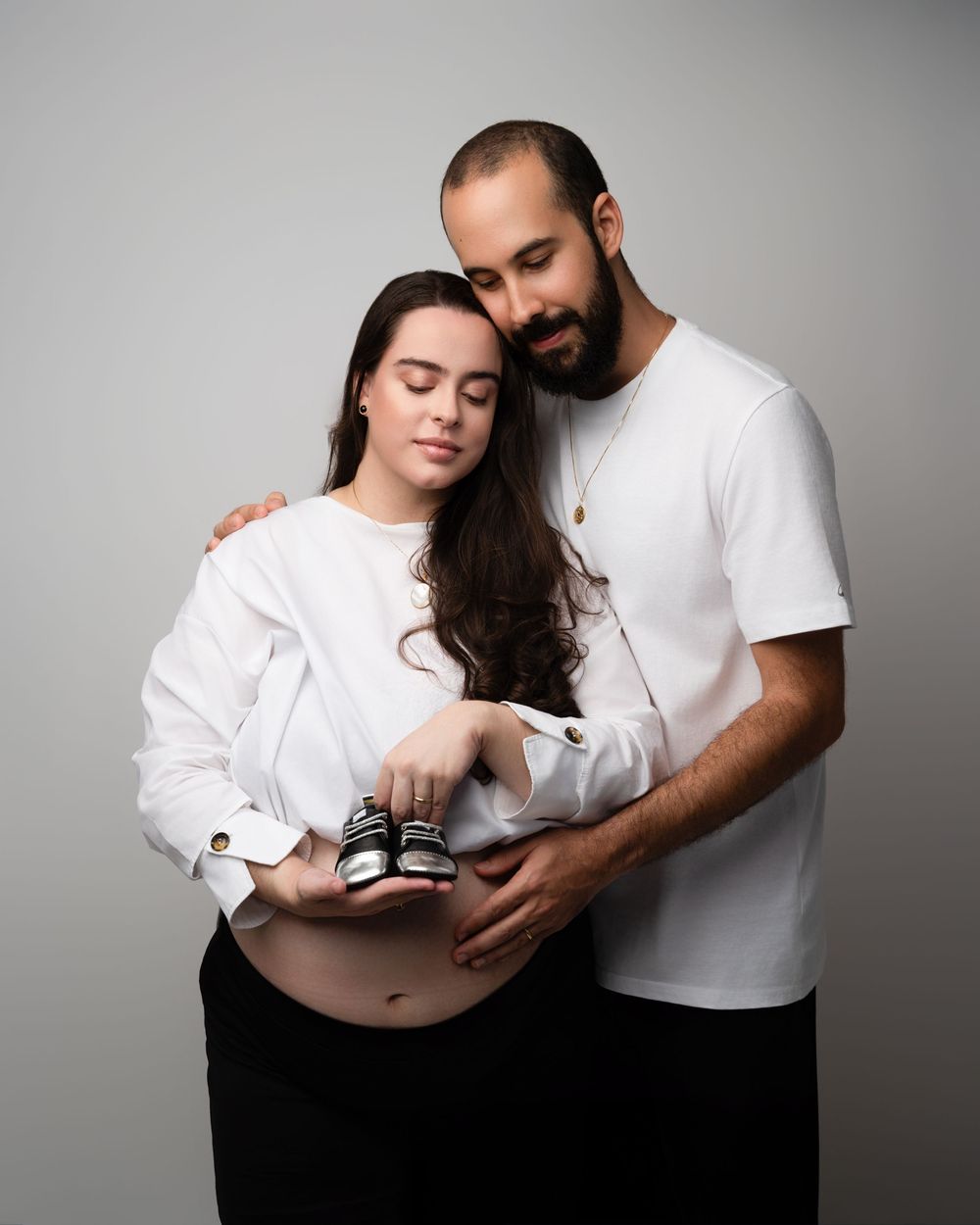 Couple in white outfits holding tiny baby shoes over pregnant belly in maternity portrait.