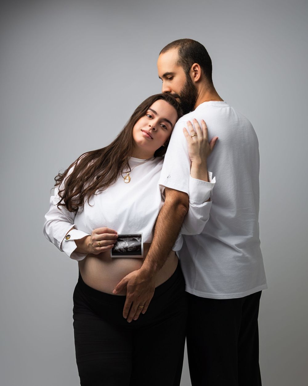 Pregnant couple in white tops holding ultrasound photo, maternity portrait on gray background.