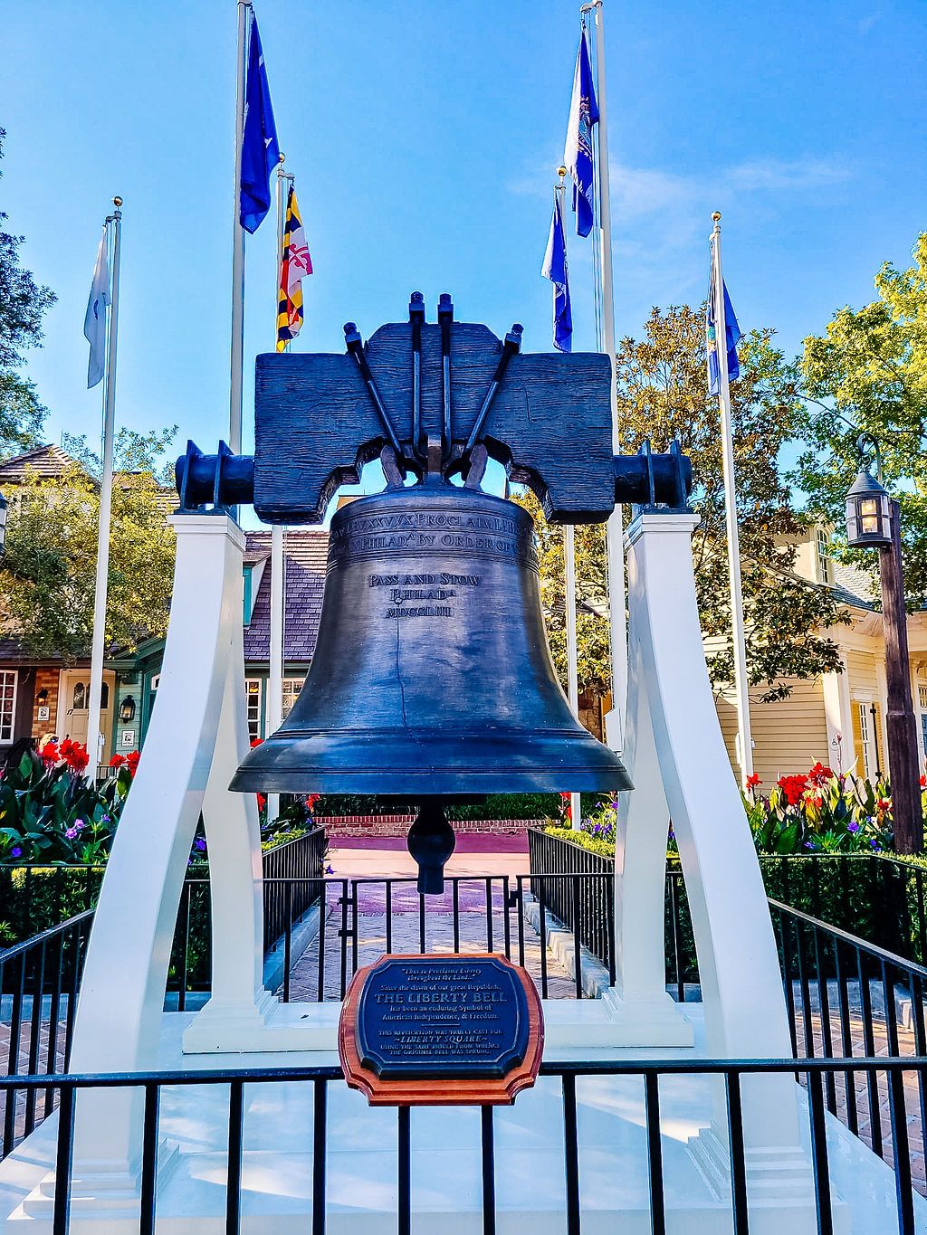 Historic Liberty Bell replica displayed outdoors with flags and white support structure against blue sky background.