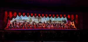 A stage performance with red curtains featuring a large ensemble cast in an ornate theatrical setting with dramatic lighting.