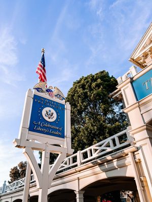 Entrance to the Liberty Square area of a theme park with American flags and white colonial-style architecture.