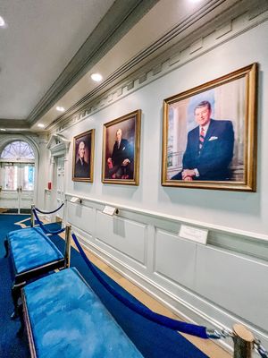 Long hallway gallery displaying portrait paintings mounted on white paneled walls with blue carpeting and recessed lighting.