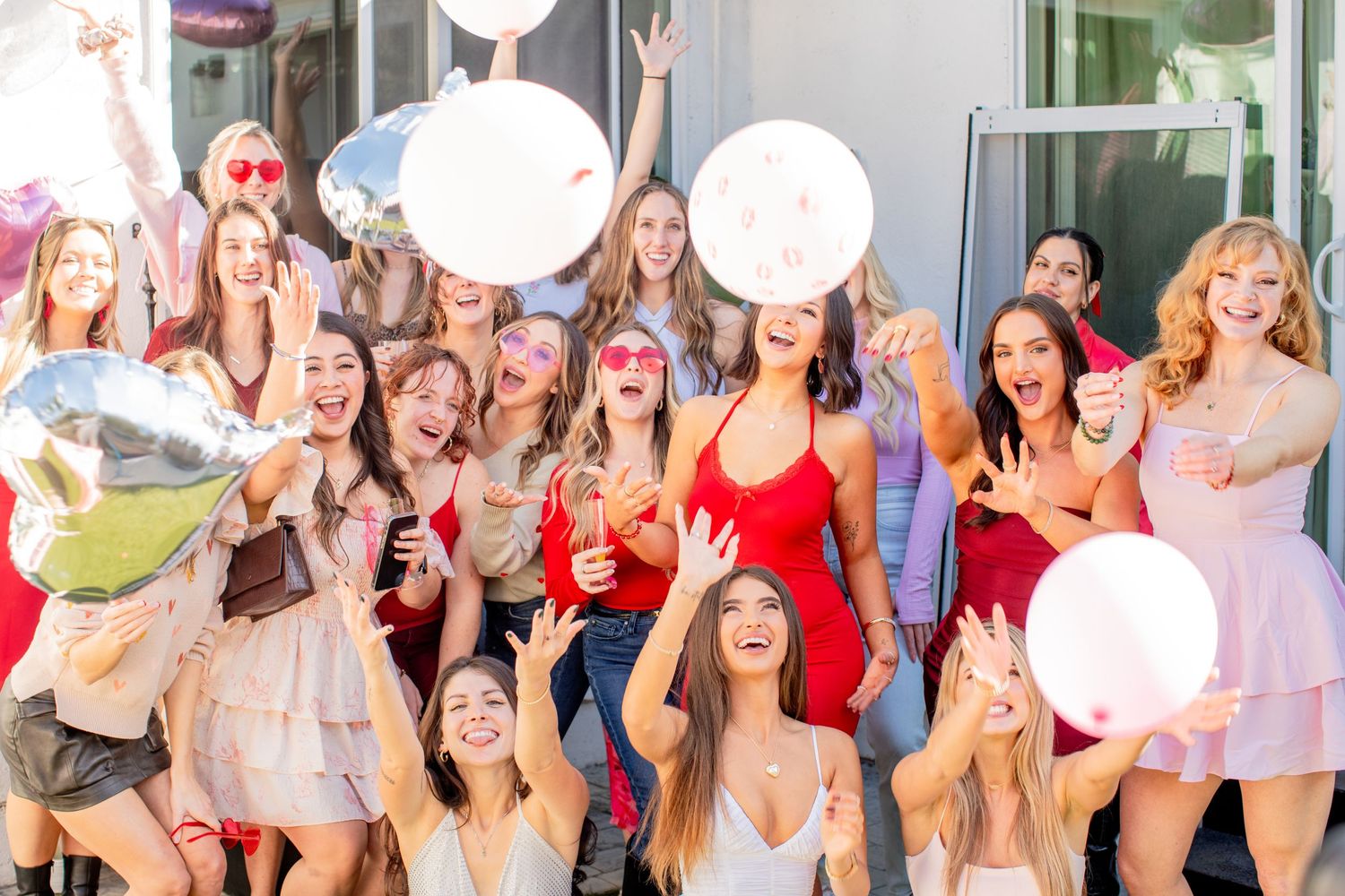 Group of white girls wearing shades of pink and red smile and cheer at the camera while tossing while tossing balloons in the air