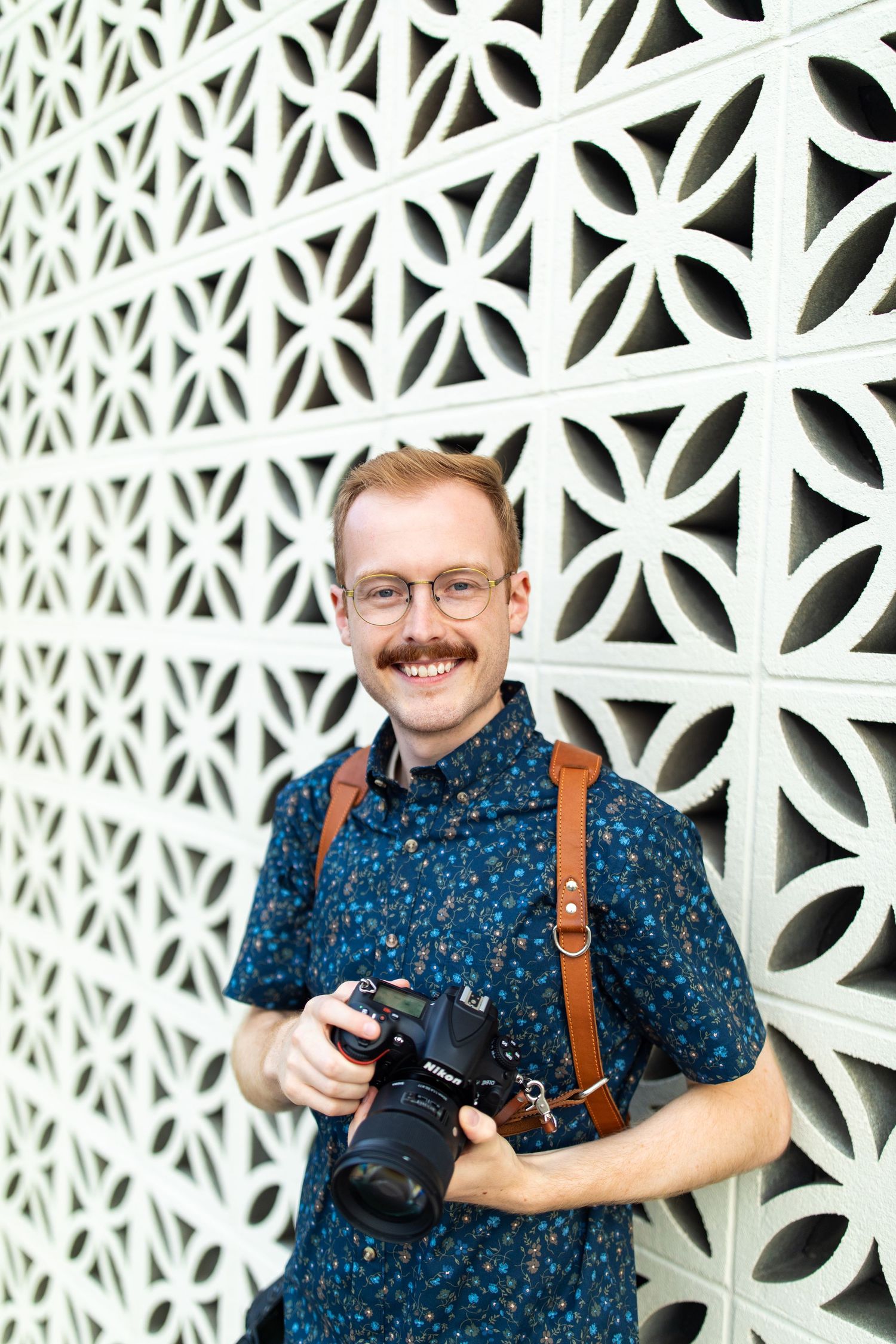 Photographer in blue floral shirt with leather camera strap smiles while holding camera against white decorative wall.