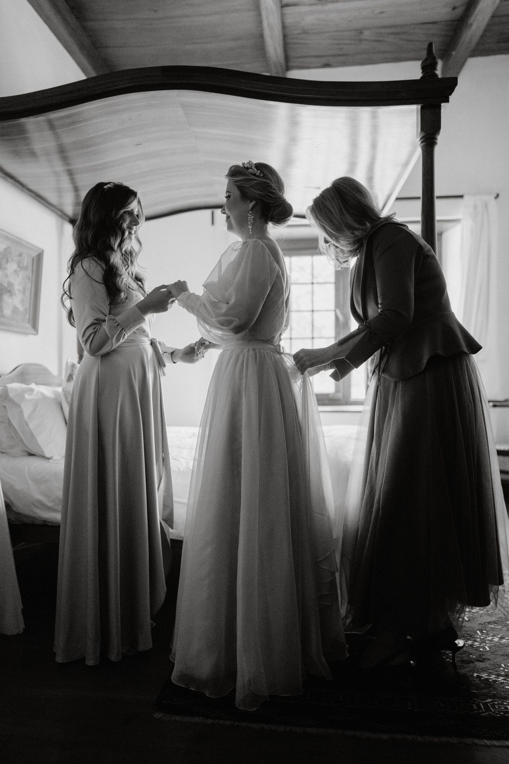 A black and white photo of three figures in elegant dresses gathered in a room with natural light streaming through windows.