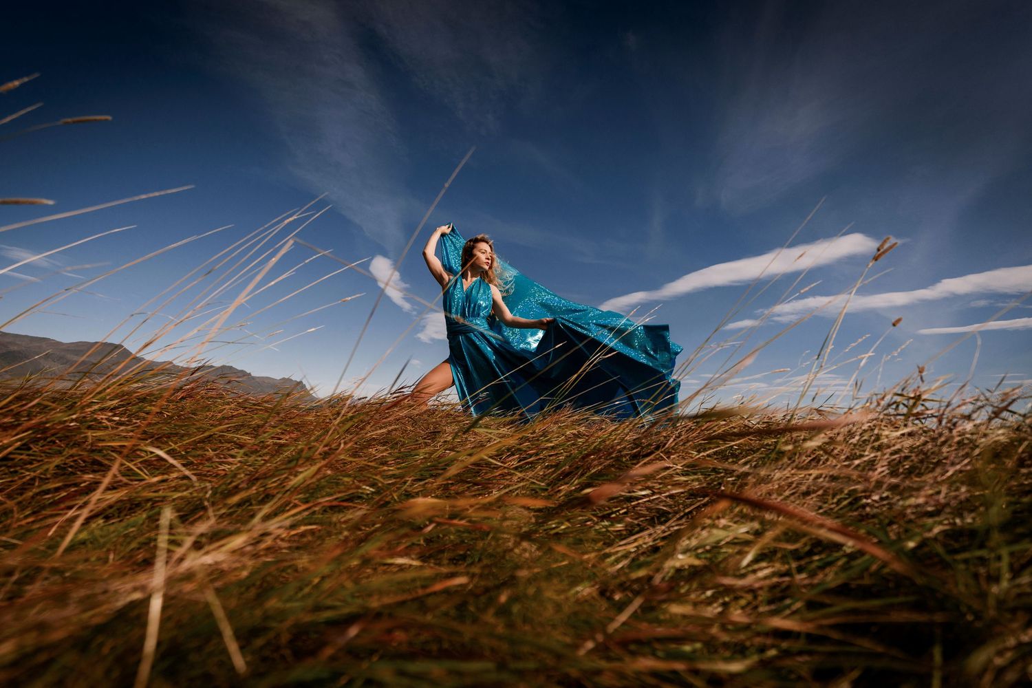 A flowing teal dress billows dramatically in the wind against a dark sky as tall grass bends in a golden field.