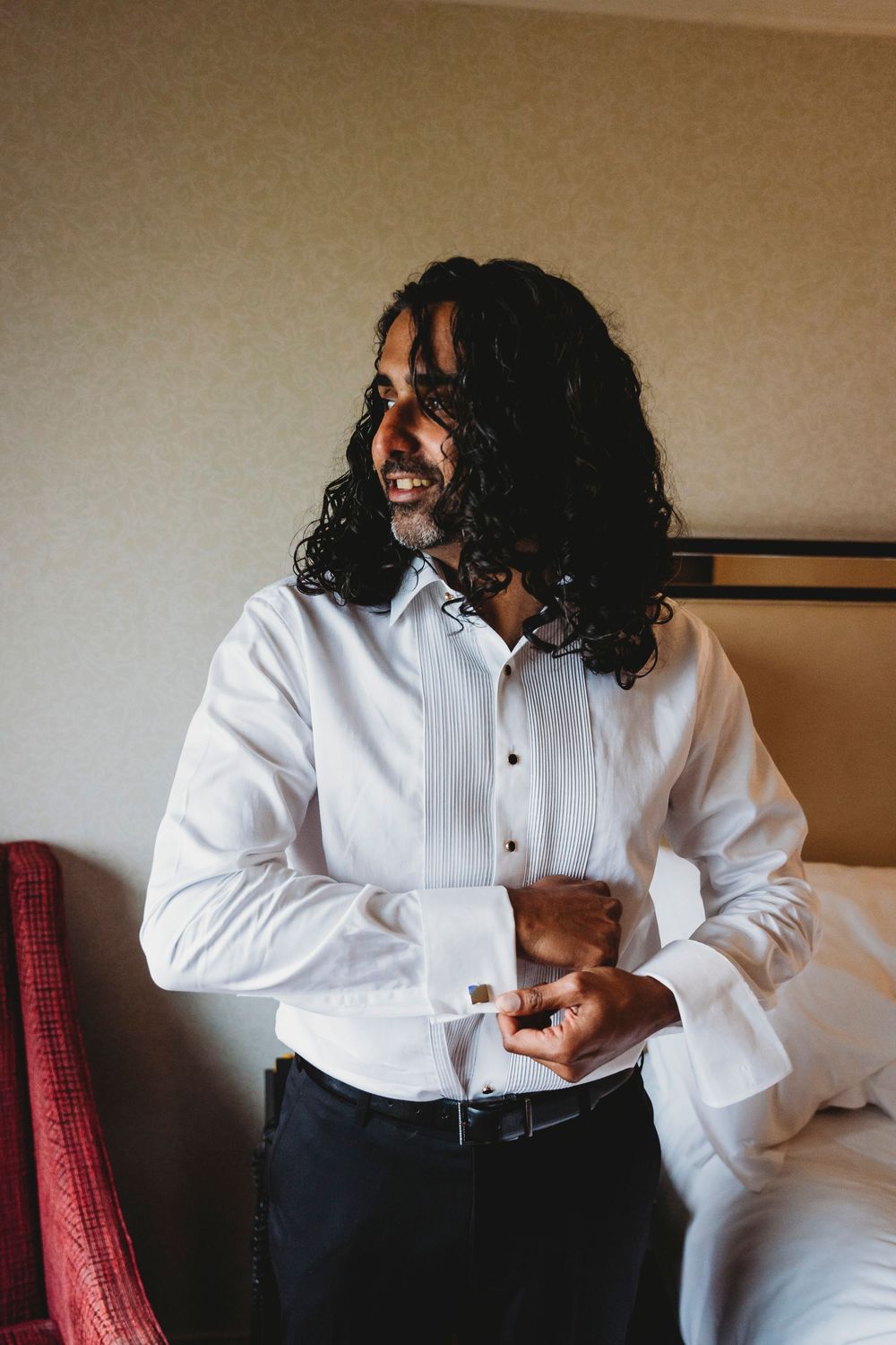 A person with long curly dark hair wearing a white dress shirt and black pants adjusts their cuff in a hotel room.