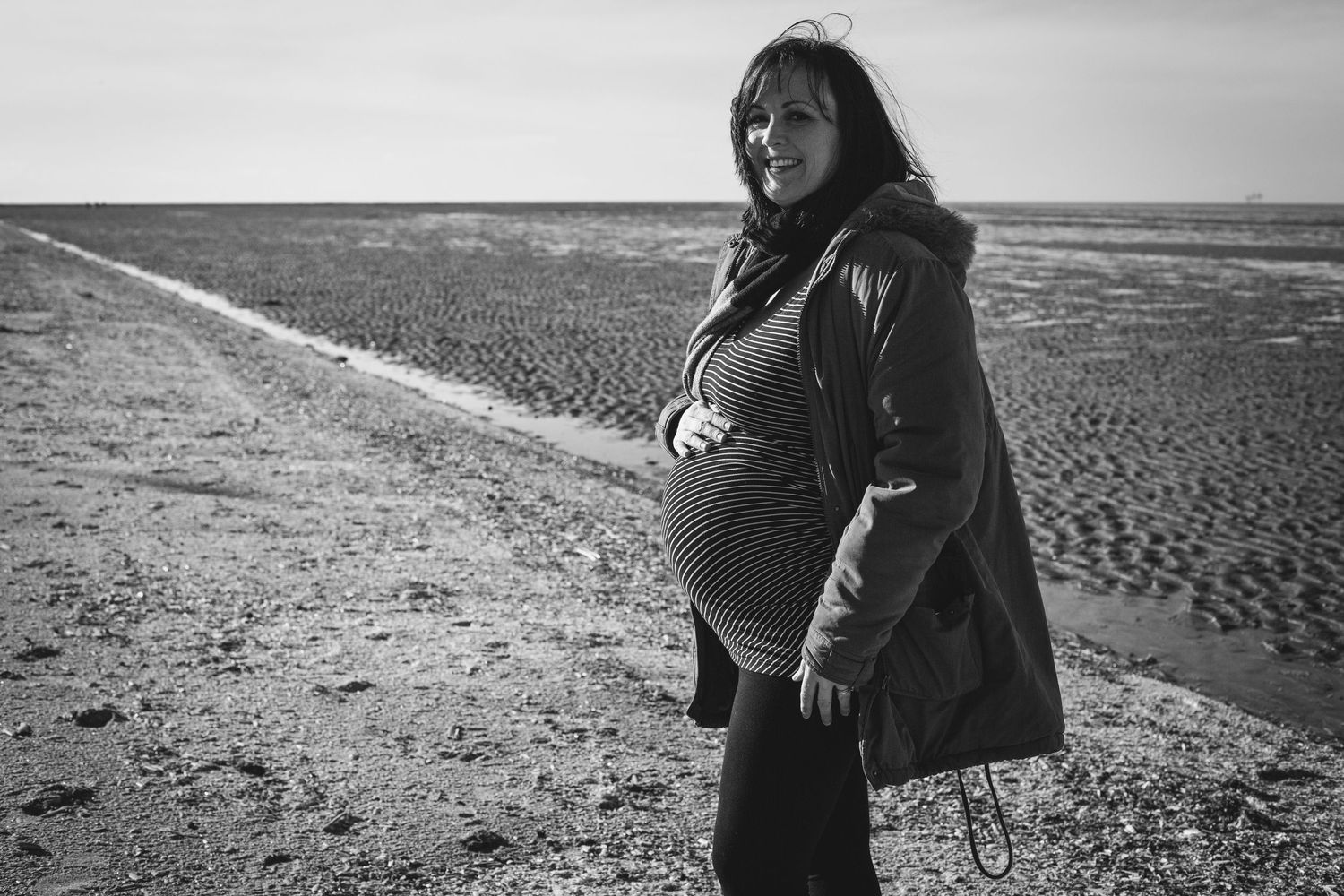 Black and white portrait of an expectant mother standing on a beach while holding her pregnant belly.