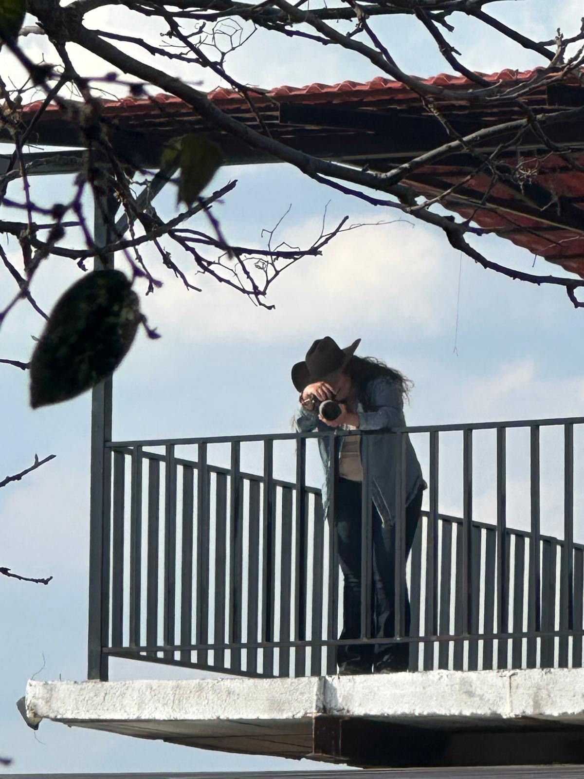 Person wearing a cowboy hat photographing from a balcony railing against a blue sky.