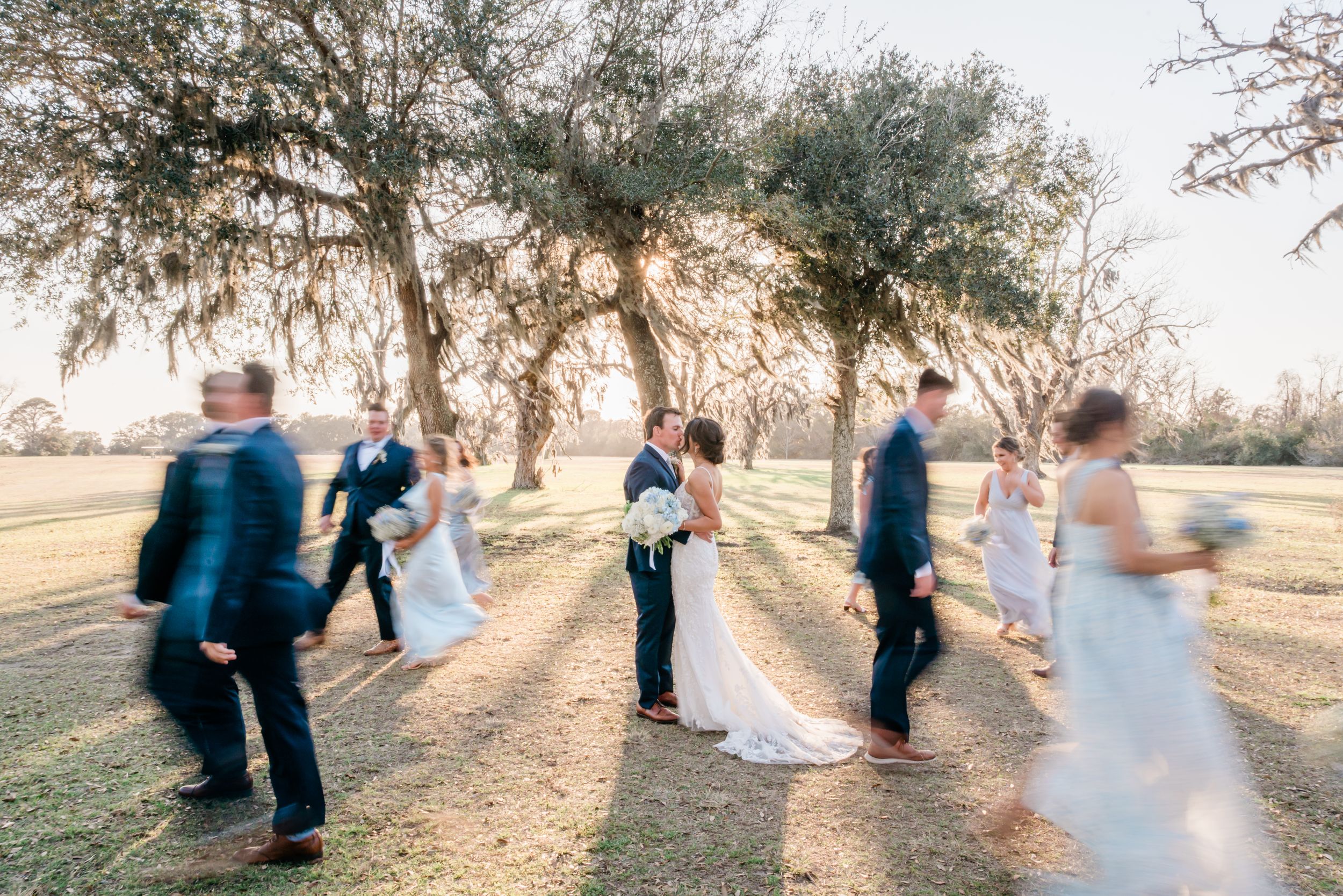 Wedding party dances in motion under sunlit trees creating dreamy silhouettes at an outdoor celebration.