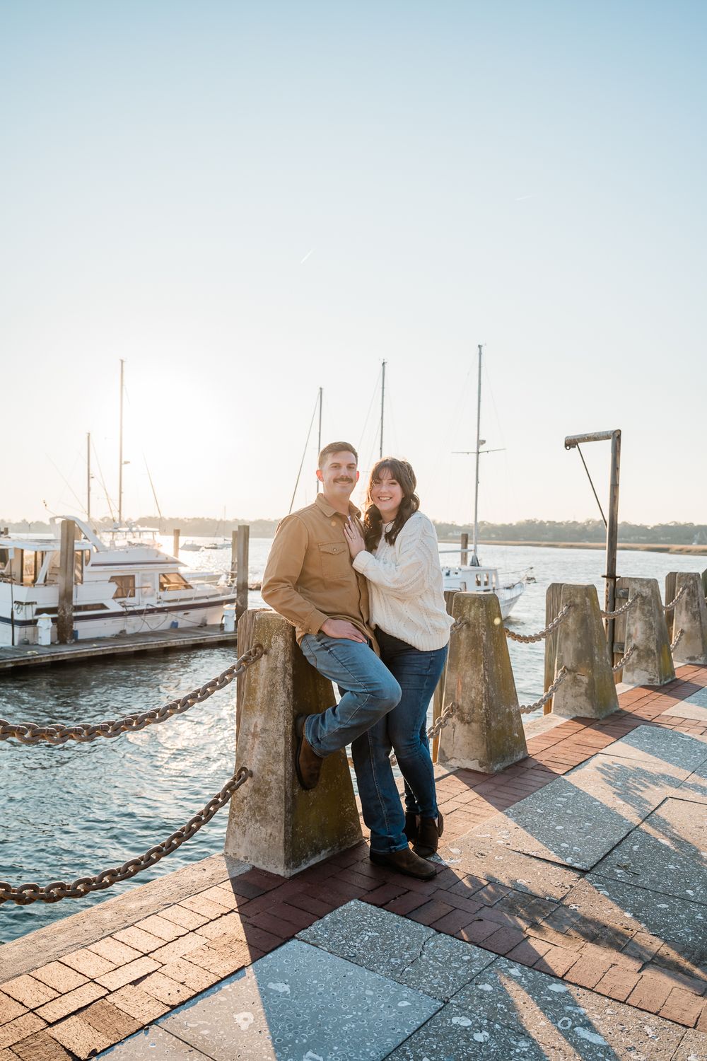 Couple posing together at marina dock with boats and water in background during sunset.