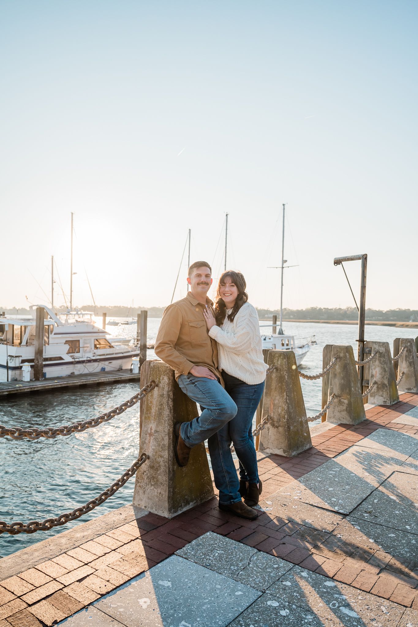 Couple posing together at marina dock with boats and water in background during sunset.