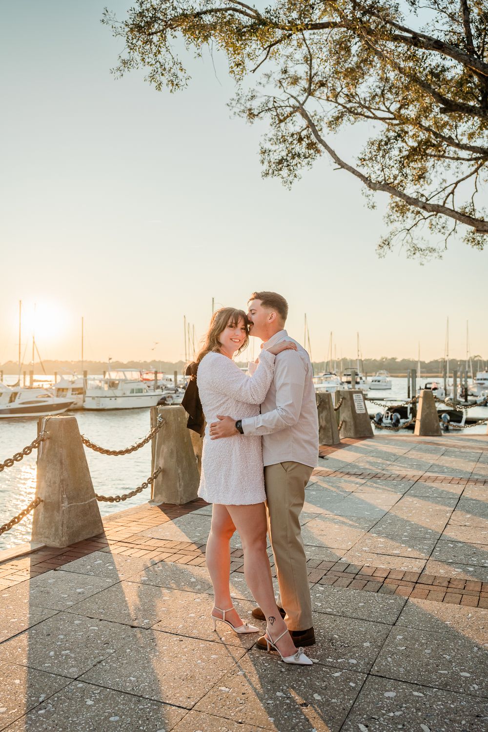 Romantic couple embracing on waterfront boardwalk during golden hour with marina in background.