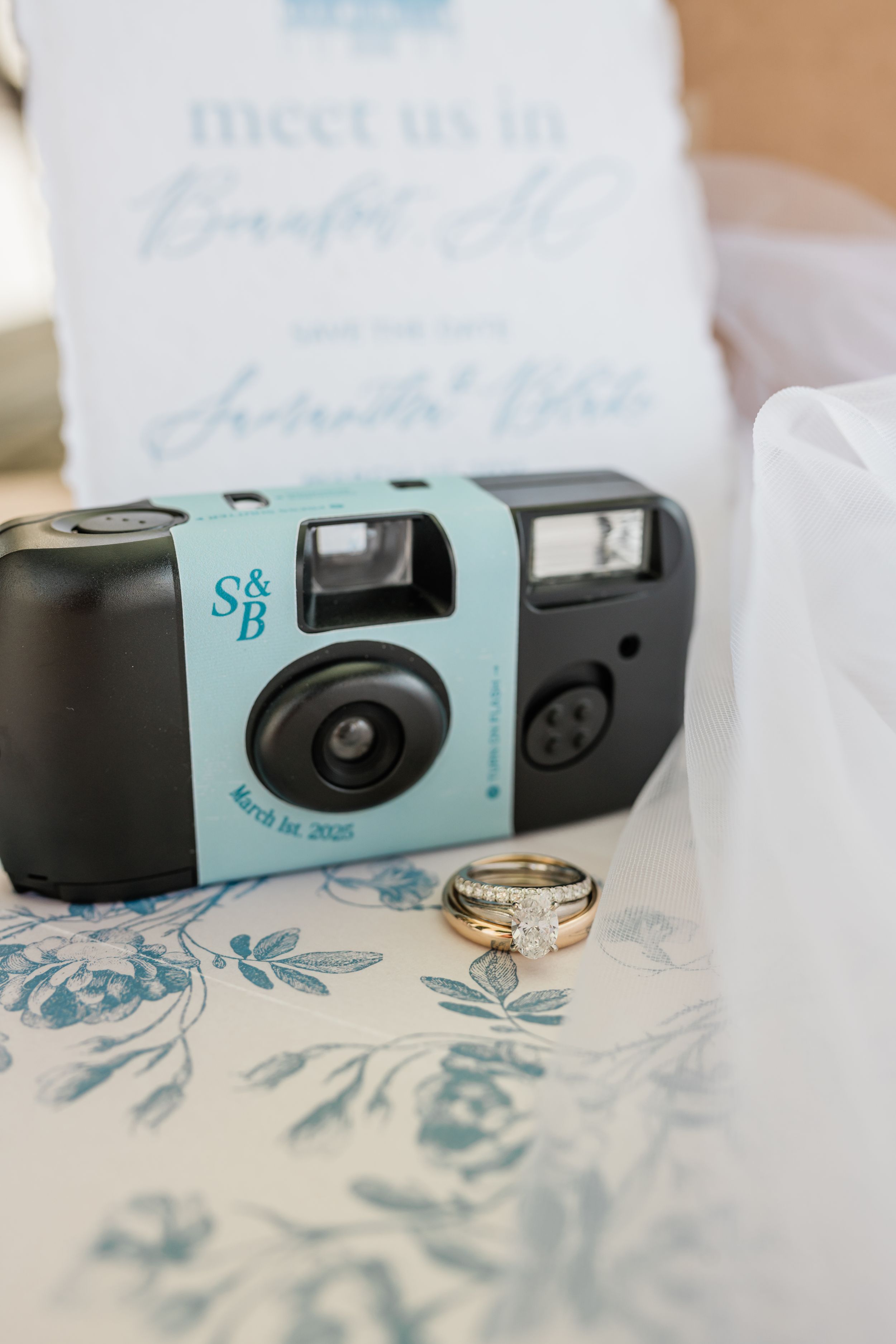Light blue disposable camera and diamond ring on floral patterned surface.