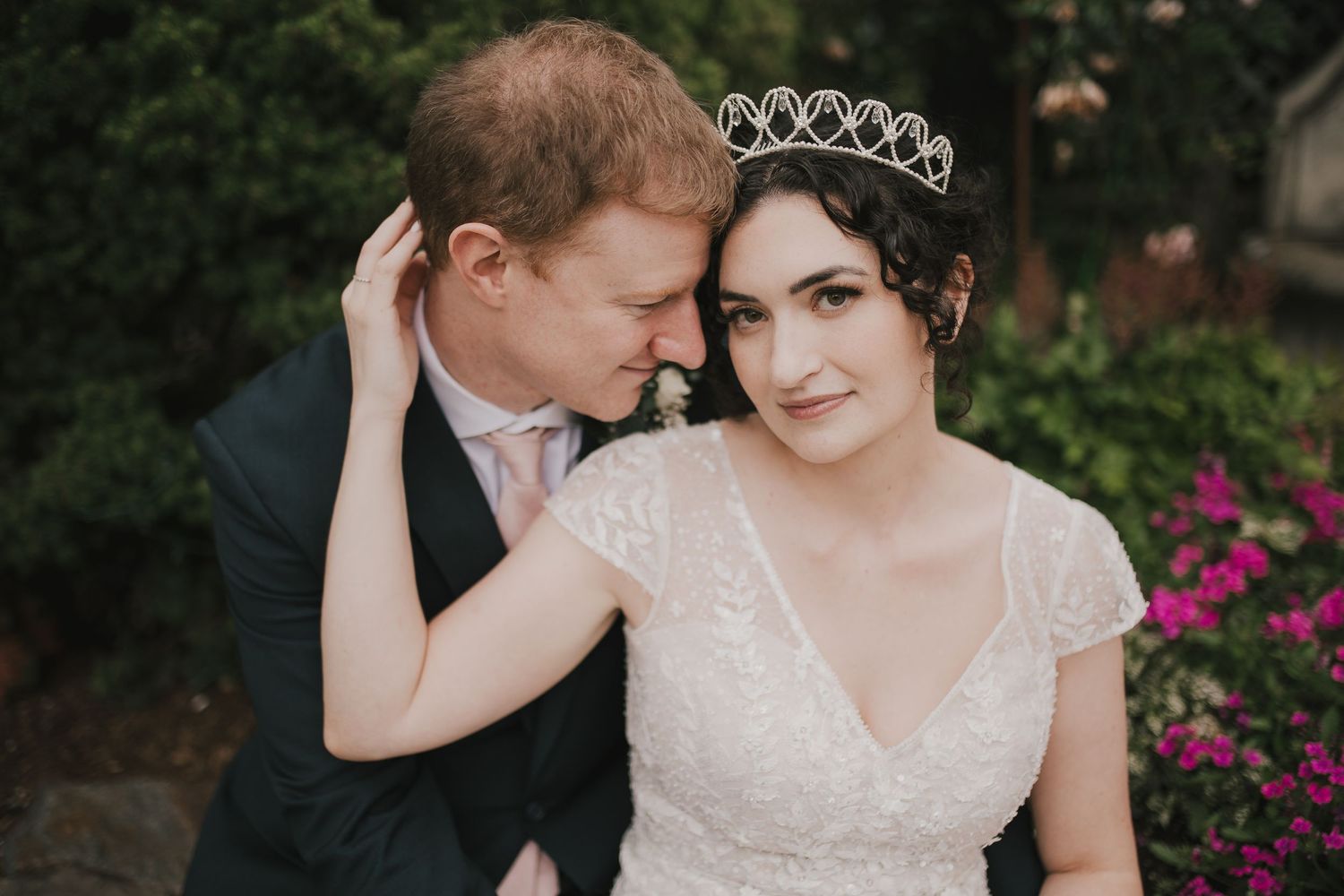 Intimate portrait of couple in wedding attire with tiara against floral garden background.