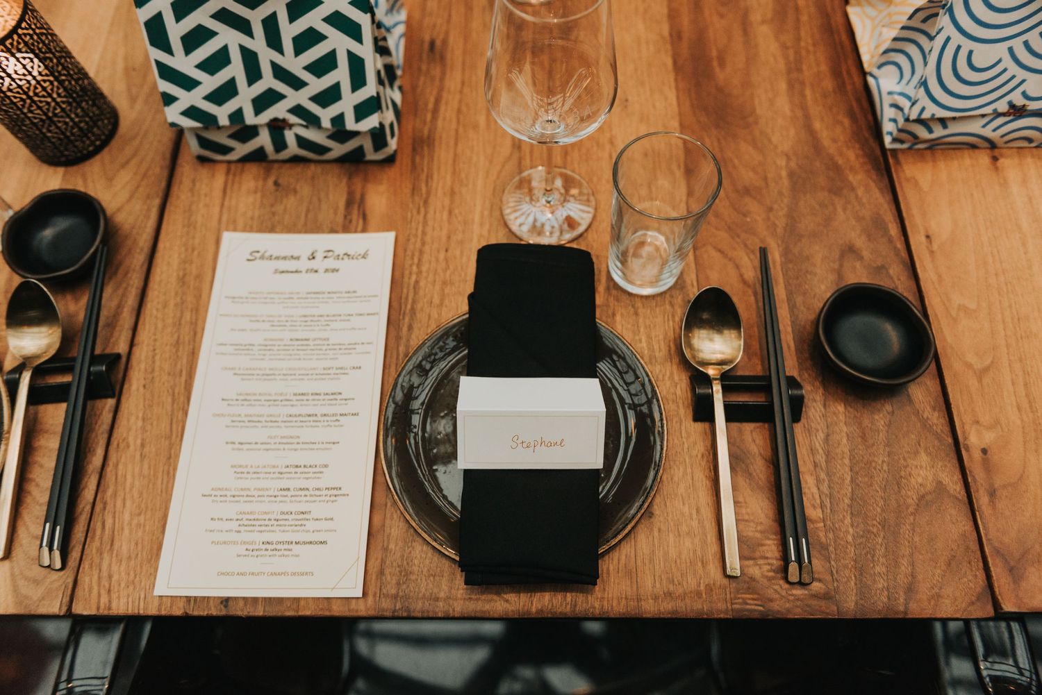 Elegant table setting with black plates, menu card, and wine glasses on wooden surface with geometric patterned pillows.