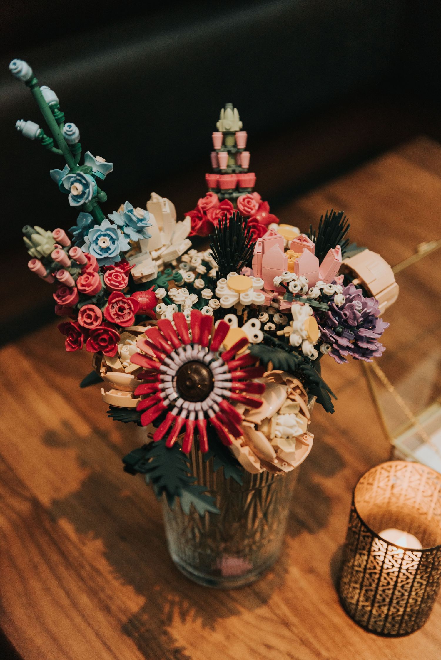 A rustic floral arrangement with red protea, blue roses, and pink flowers in a glass jar beside a decorative candle holder.
