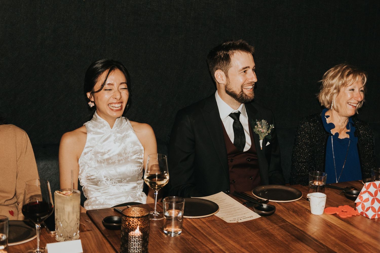 People enjoying a wedding reception at a long dining table with drinks and dim lighting.