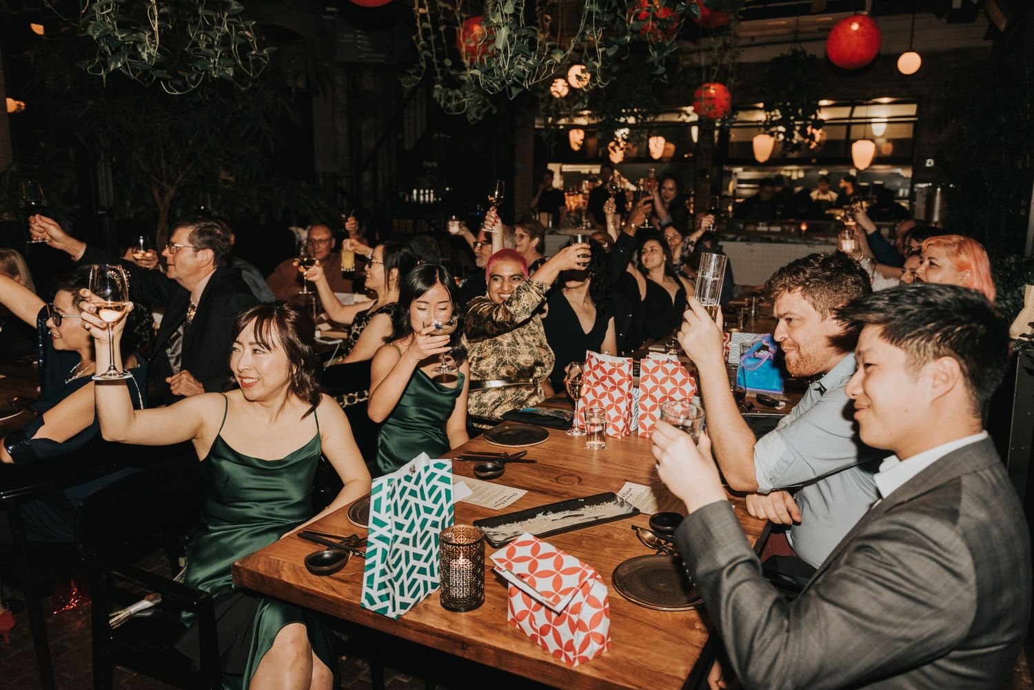 Crowded bar patrons raise their hands and cheer during a festive nighttime celebration illuminated by red lights.