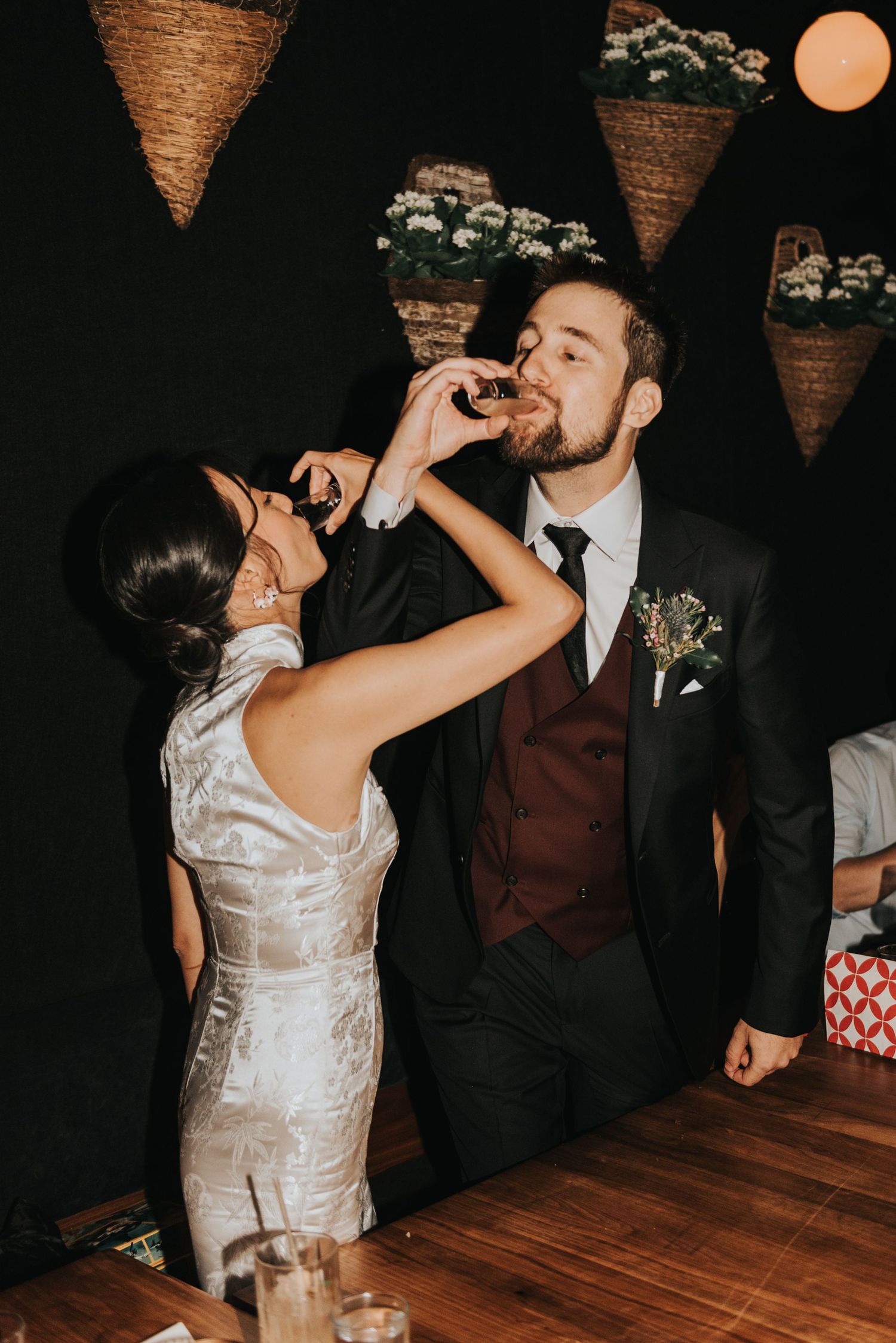 Wedding couple feeding each other at their reception with hanging floral decor and wooden floor below.