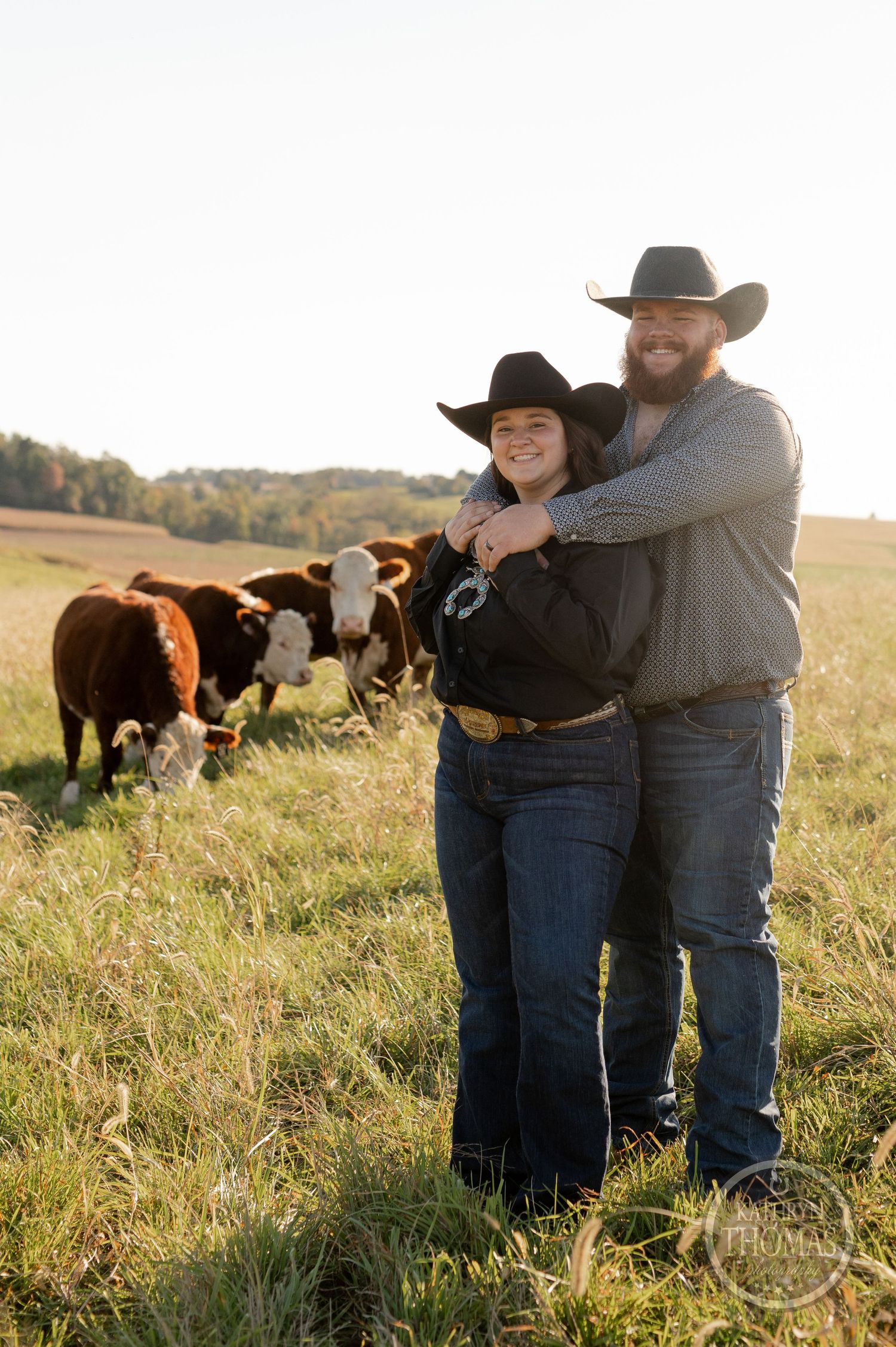 Couple in western wear embrace on their ranch with Hereford cattle grazing in the pasture behind them.