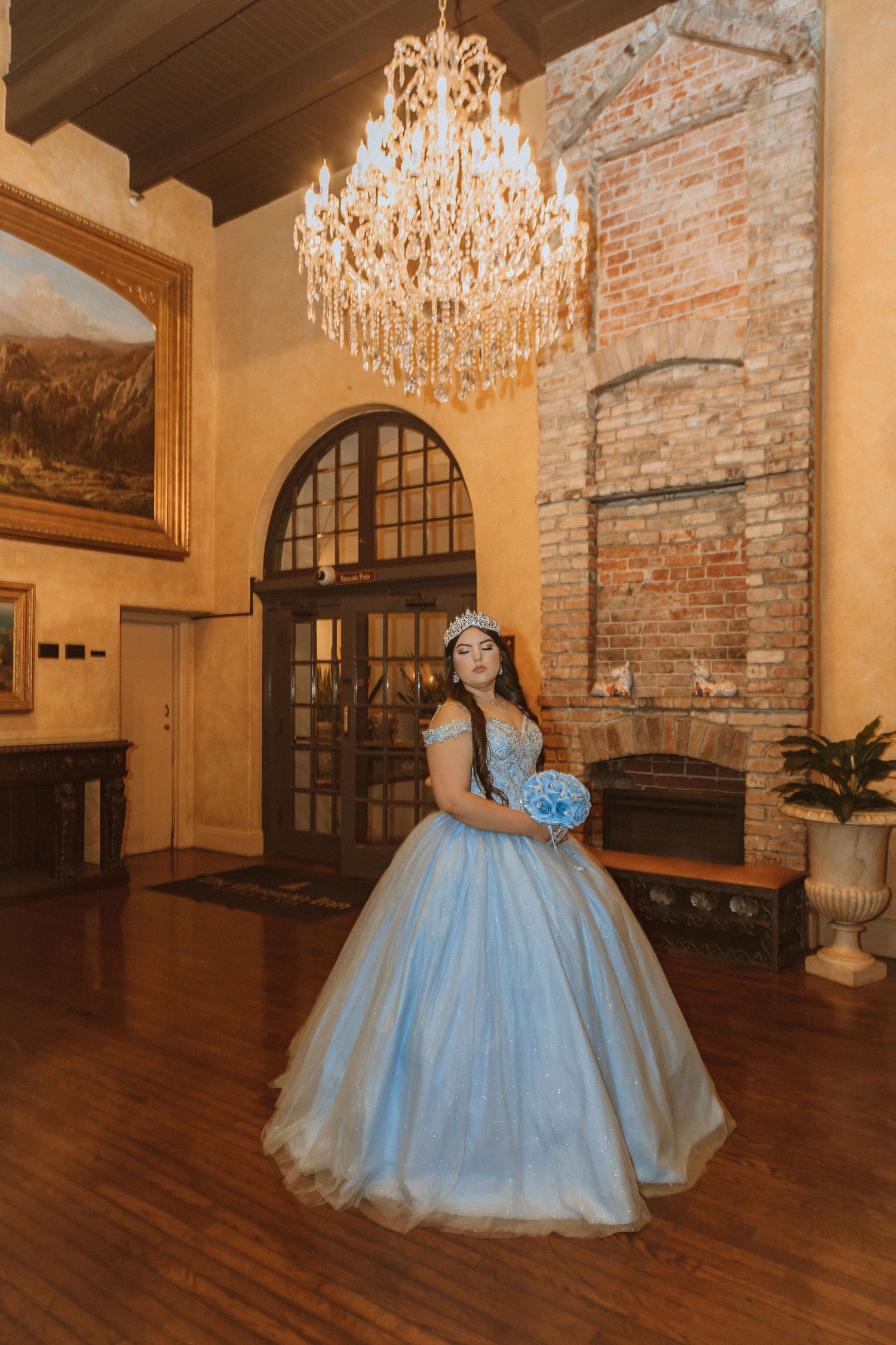 A stunning blue ballgown sparkles beneath an elegant crystal chandelier in a room with exposed brick and hardwood floors.