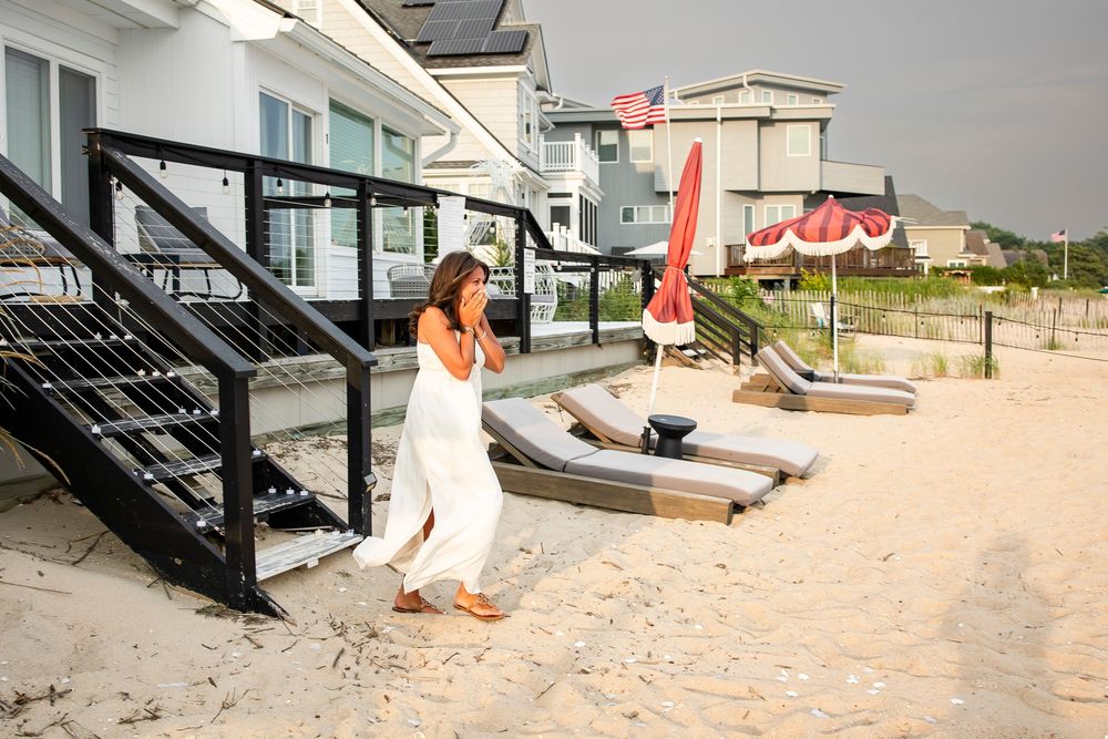 Two people in white dresses stand near lounge chairs on a sandy beach by modern beach house steps.
