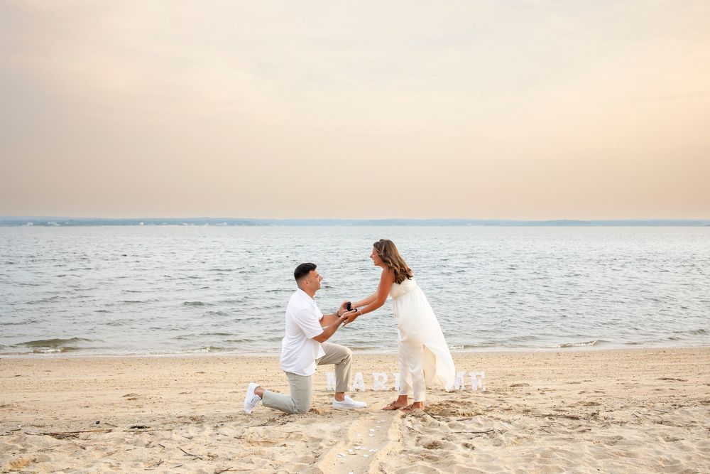 A couple in white outfits shares a romantic moment on a sandy beach at sunset by the ocean.