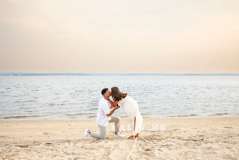 A person in white clothing sits on a sandy beach near the ocean during sunset, creating a peaceful coastal scene.
