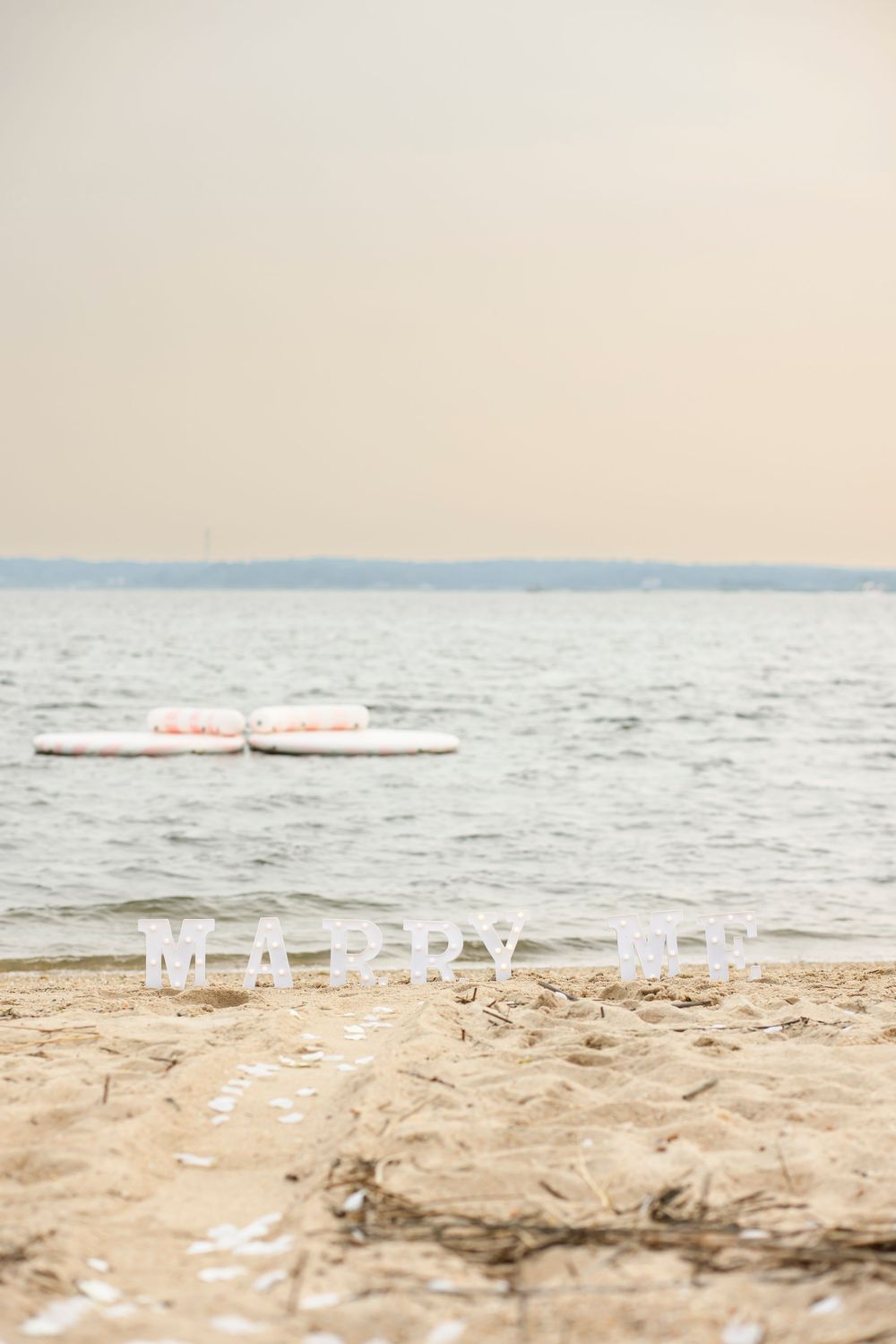 White 'MARRY ME' letters arranged on a sandy beach with ocean waves in the background during sunset.